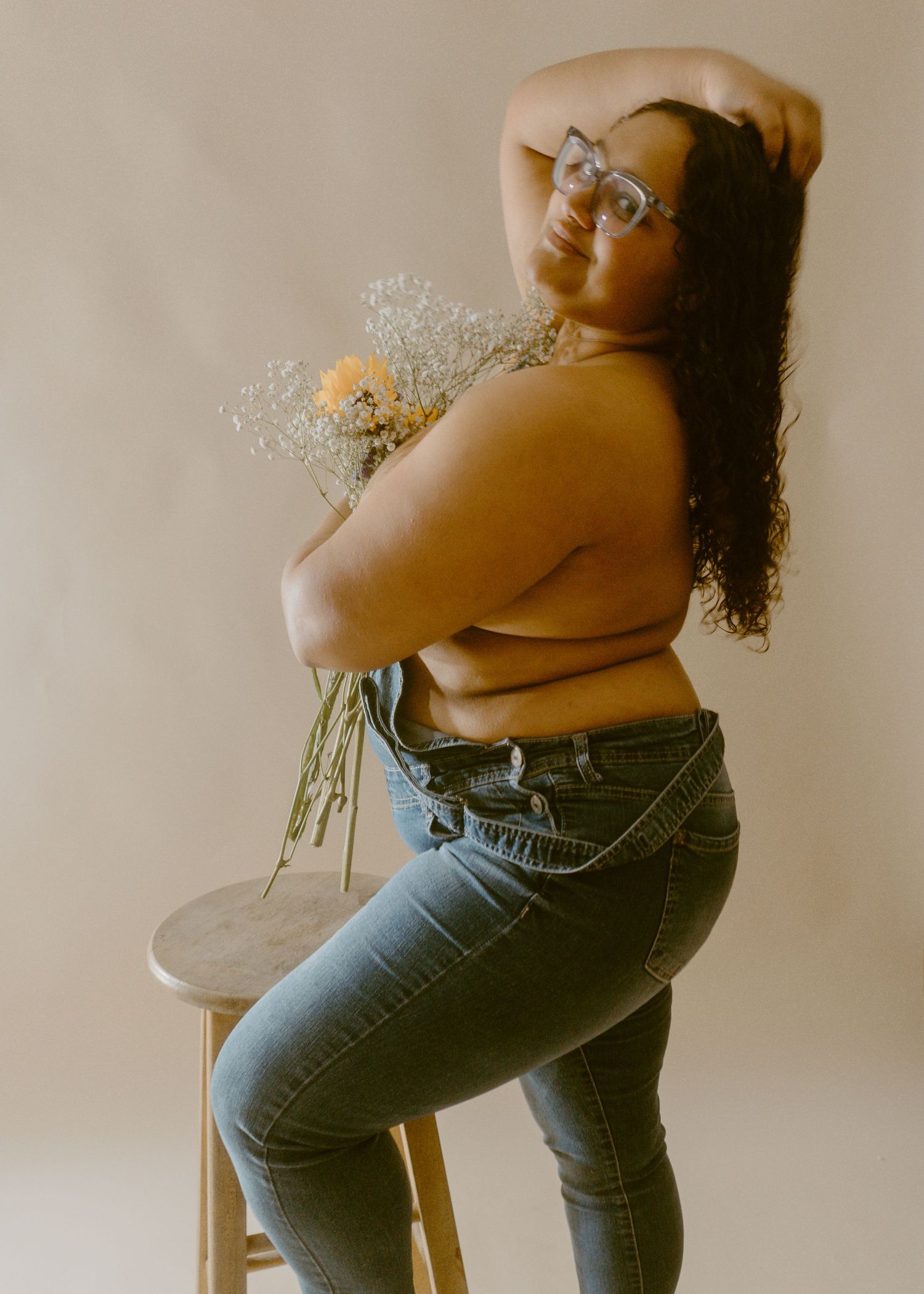 Woman in jeans holding wildflowers, posing on stool in studio, wearing glasses with curly hair.