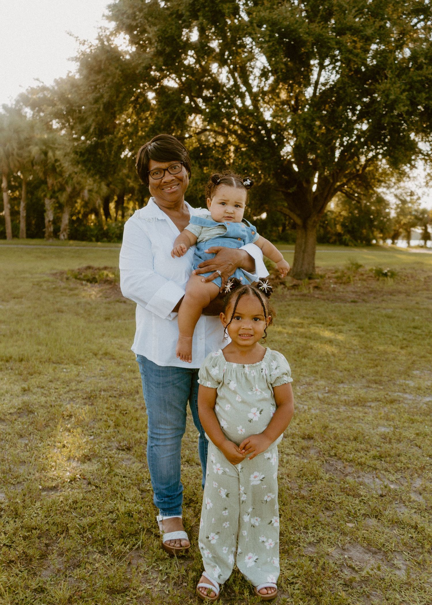 A woman holding a baby stands with a young girl in a sunny park, surrounded by lush green trees.