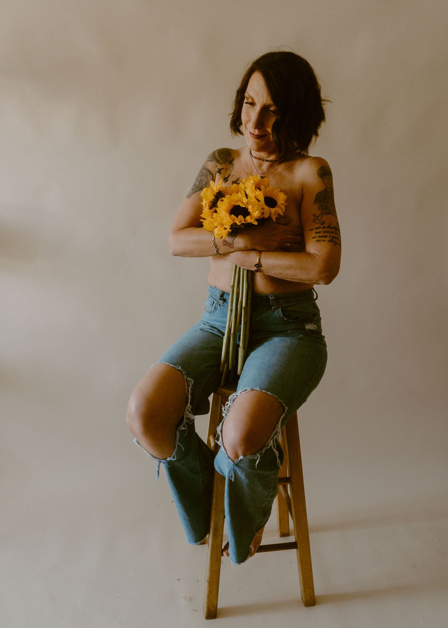 Tattooed woman sitting on stool holding yellow sunflowers, wearing ripped jeans, smiling softly.