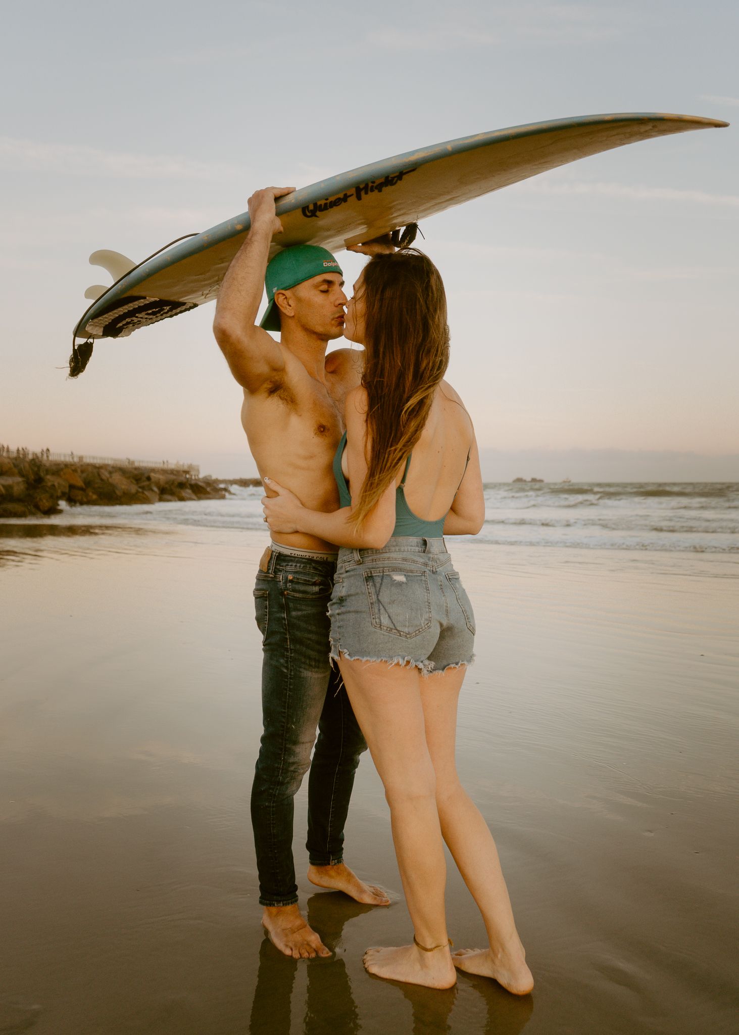 Couple kissing on beach at sunset, man holding surfboard overhead, woman in denim shorts embracing shirtless surfer.