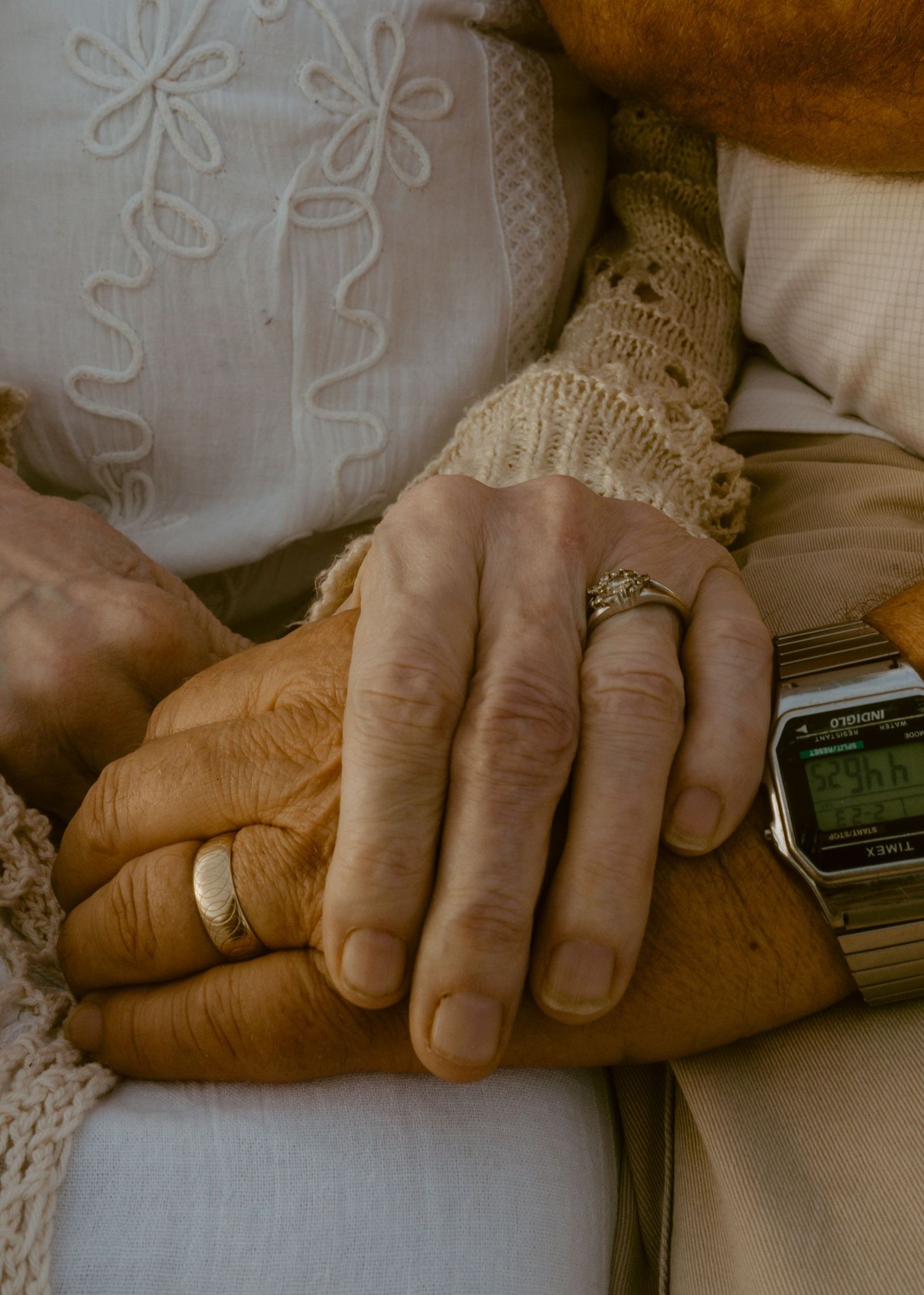 Elderly couple holding hands, wearing wedding rings and a vintage watch, dressed in lace and embroidered clothing.