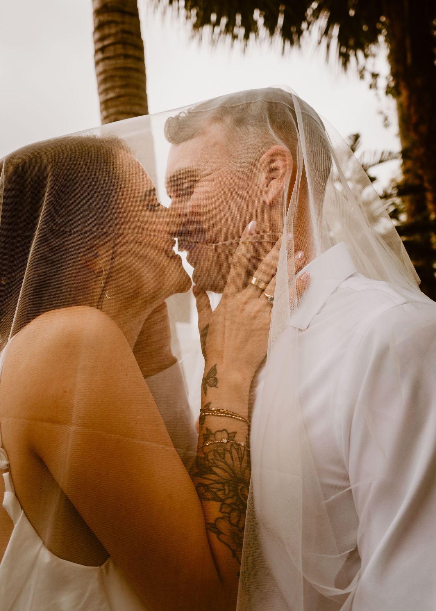 Bride and groom sharing a romantic kiss under a wedding veil, surrounded by palm trees on their tropical wedding day.