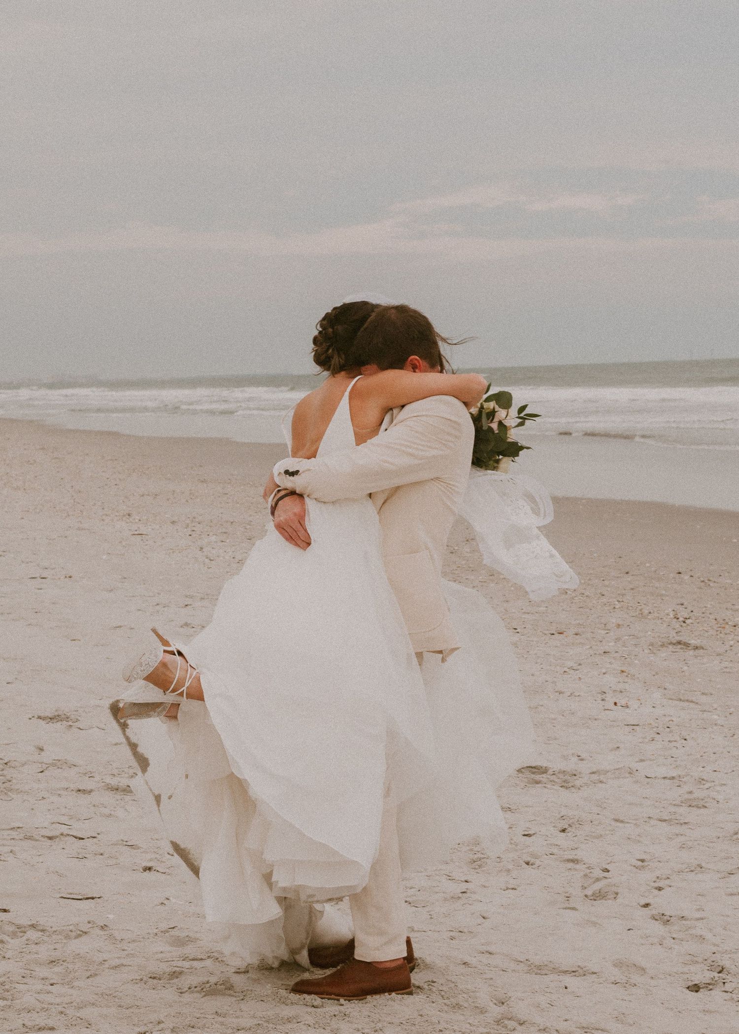 Bride and groom embrace on sandy beach, bride in white gown holding bouquet.