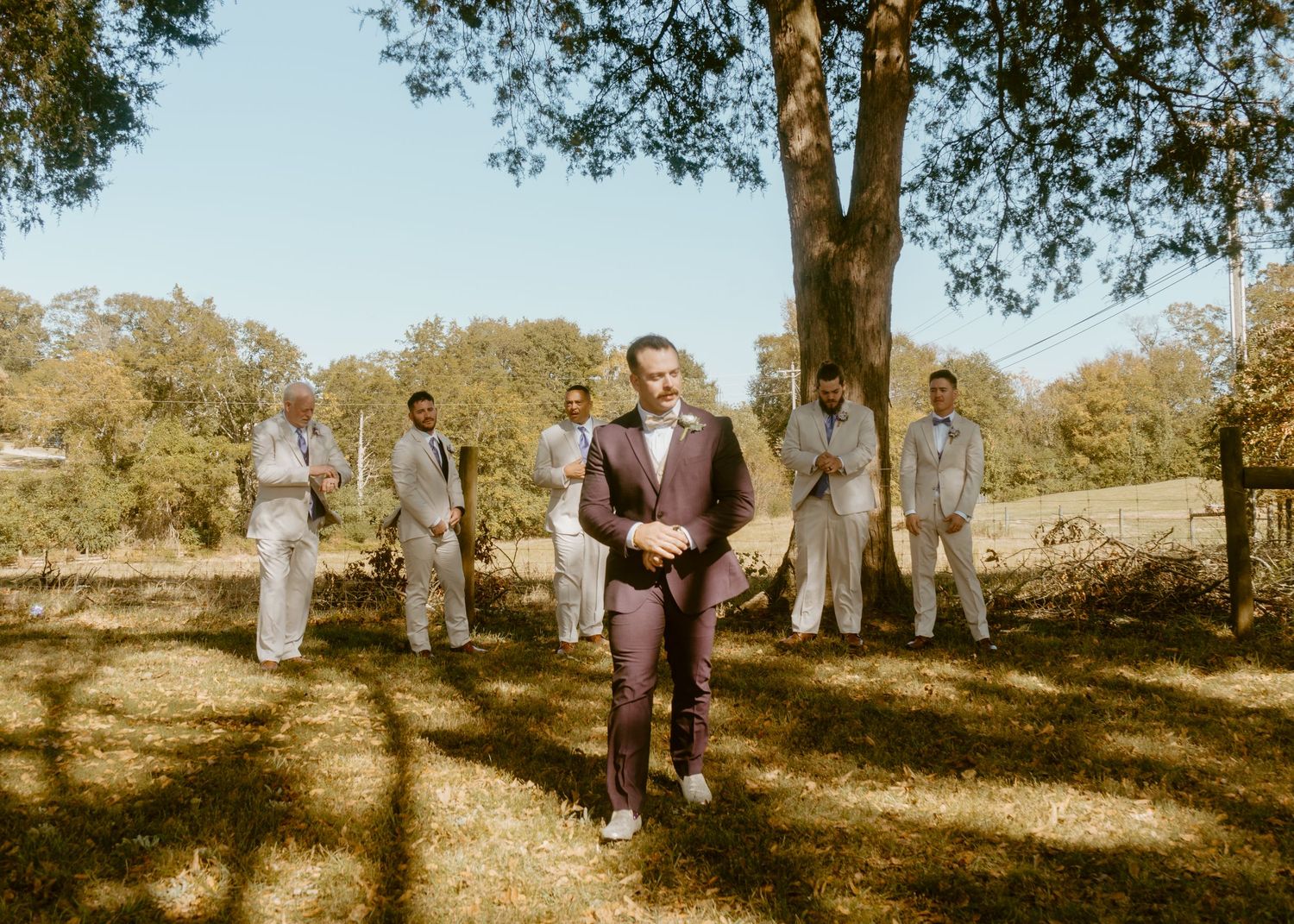 Groom in burgundy suit stands confidently before groomsmen in gray suits outdoors under trees on a sunny wedding day.