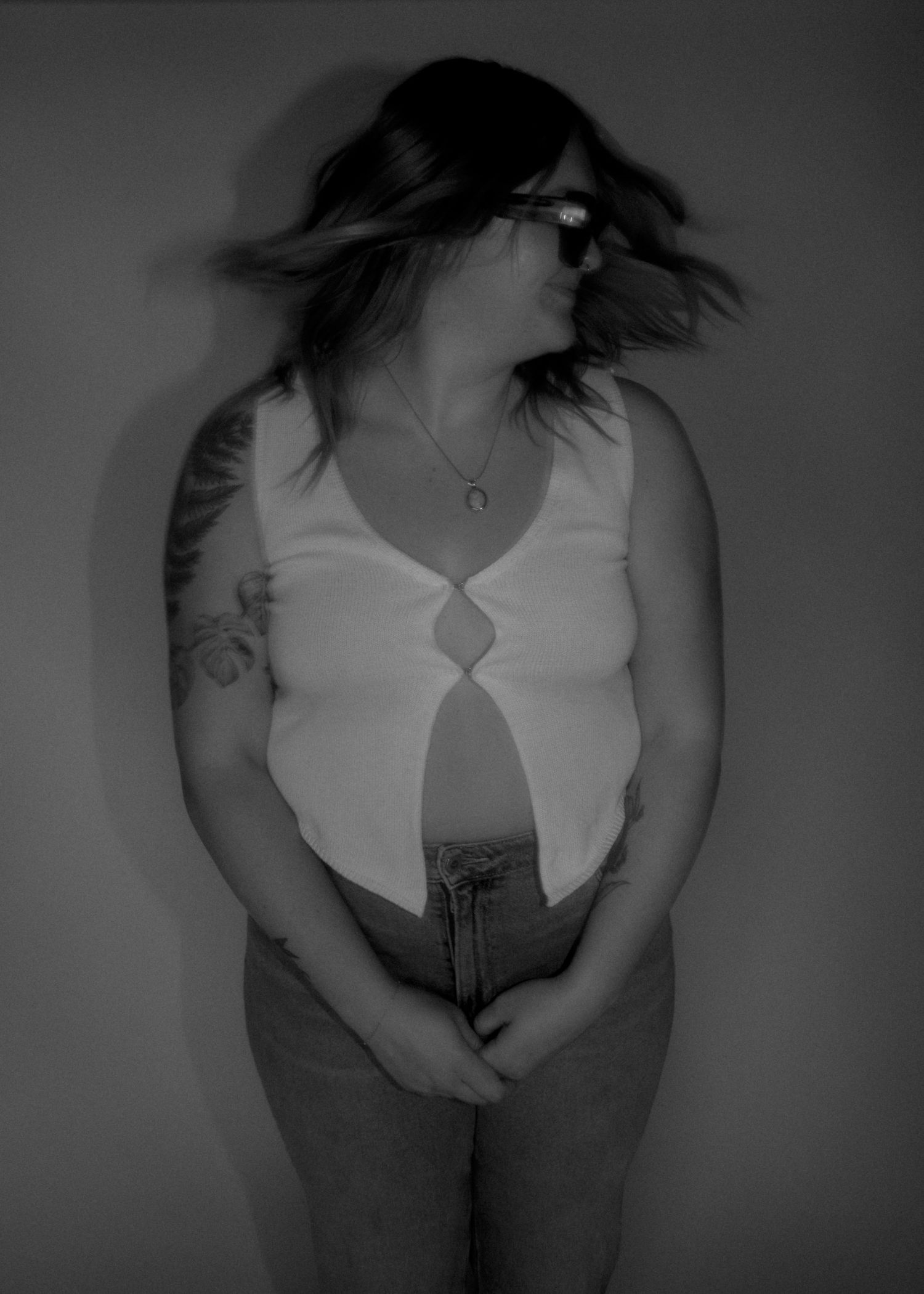 Black and white photo of a woman with tattoos, glasses, and a sleeveless top, hair in motion.