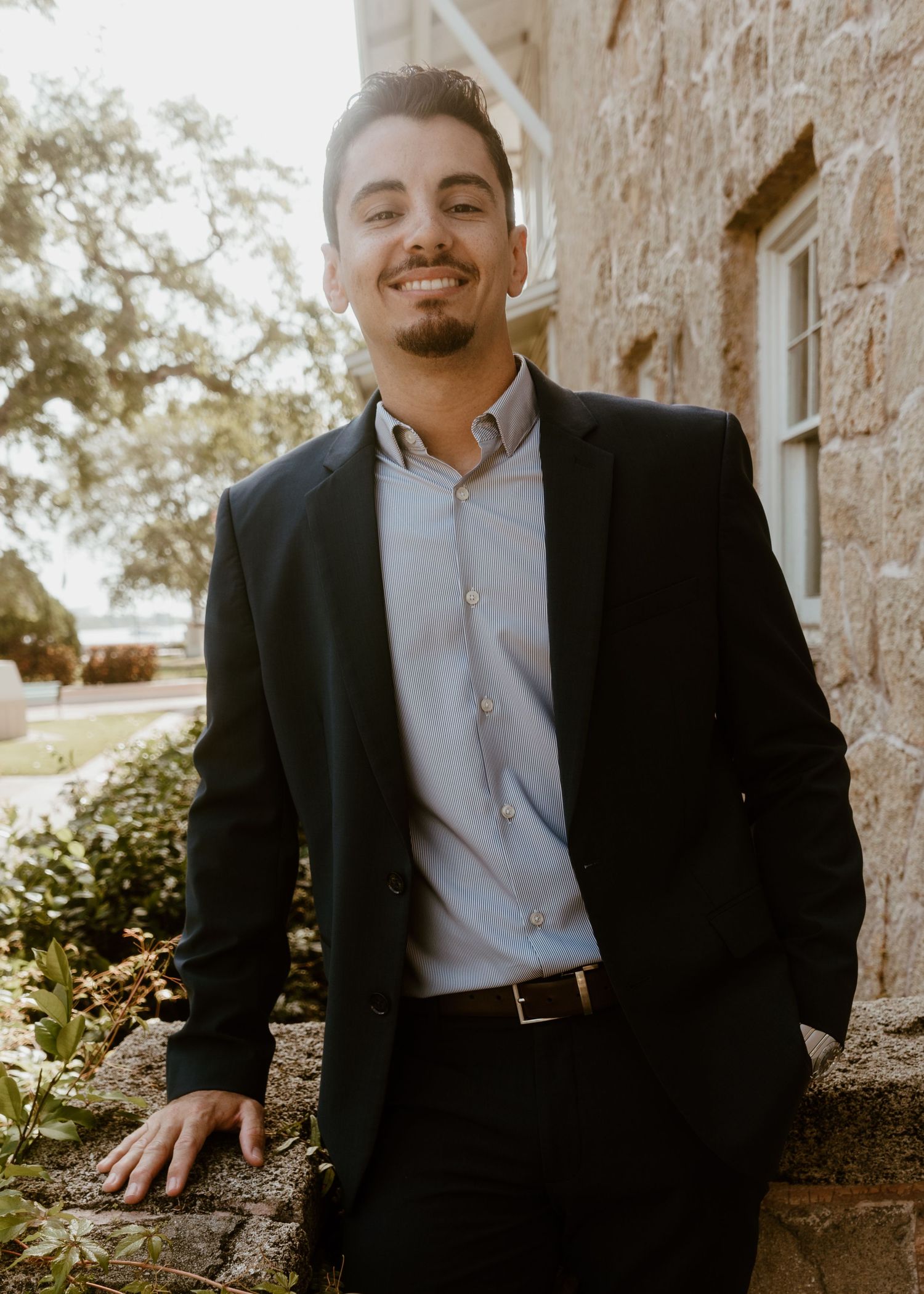 Young man in black blazer and striped shirt smiling outdoors near stone building.