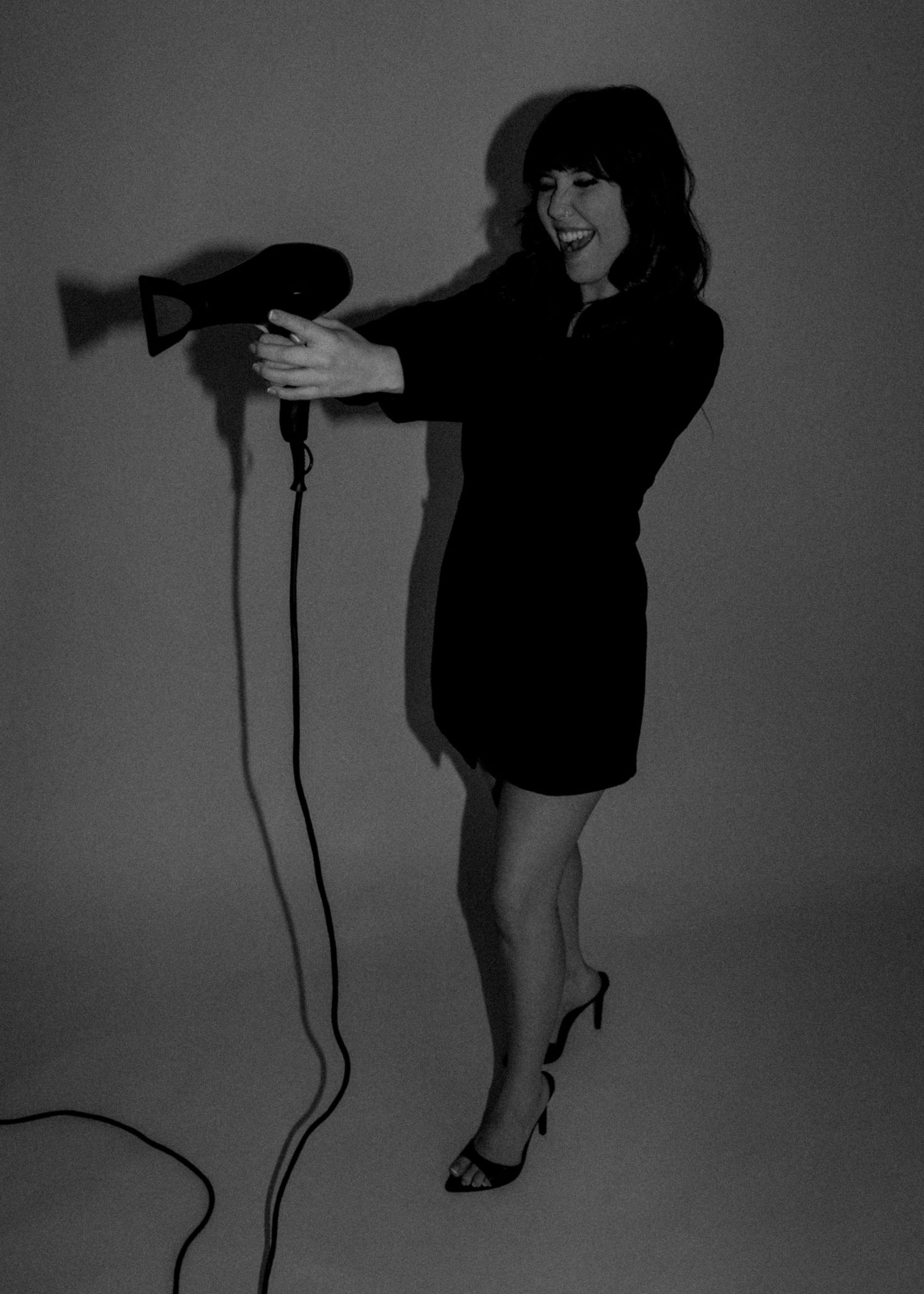 Woman in black dress holding a hair dryer, posing playfully in a black and white studio photo.