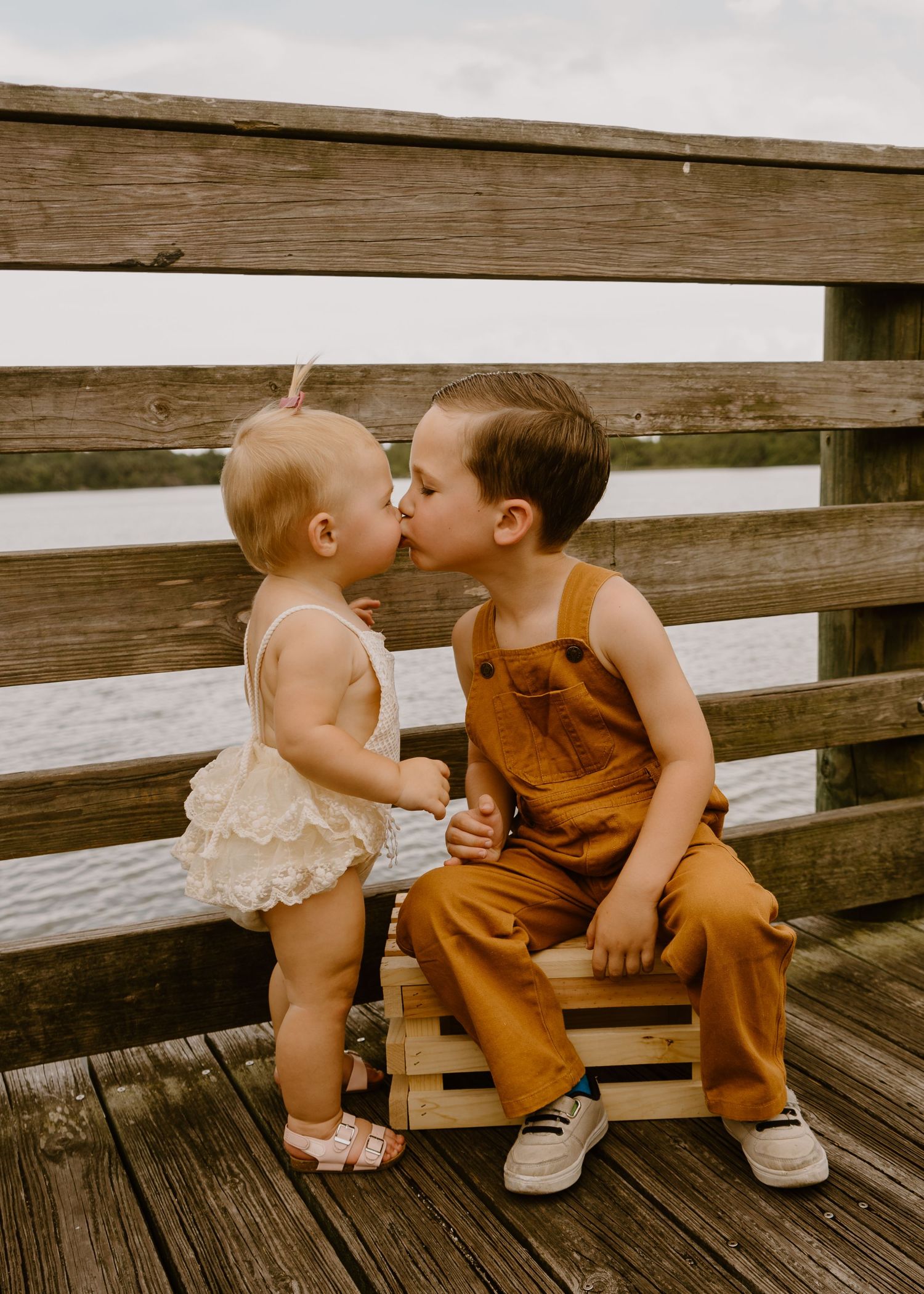 Two toddlers share a sweet kiss on a wooden pier, boy in brown overalls and girl in white ruffled outfit.