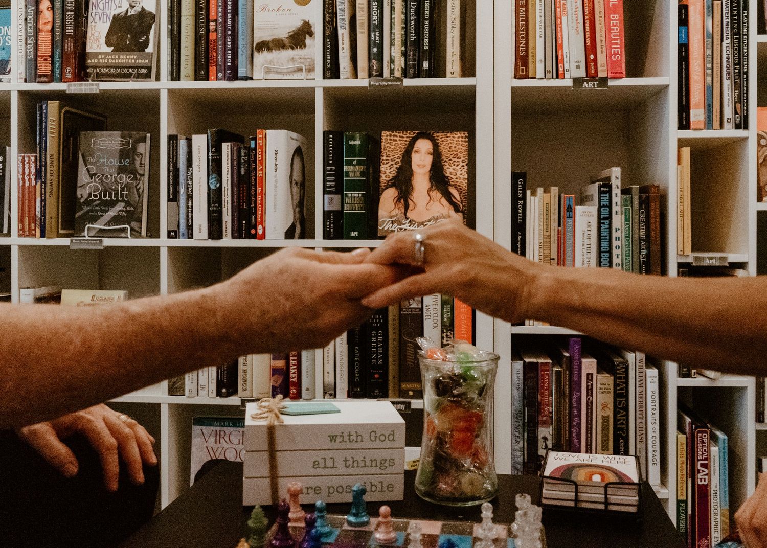 Two hands exchanging a ring over a table with books and a framed photo in a cozy bookshelf-lined room.