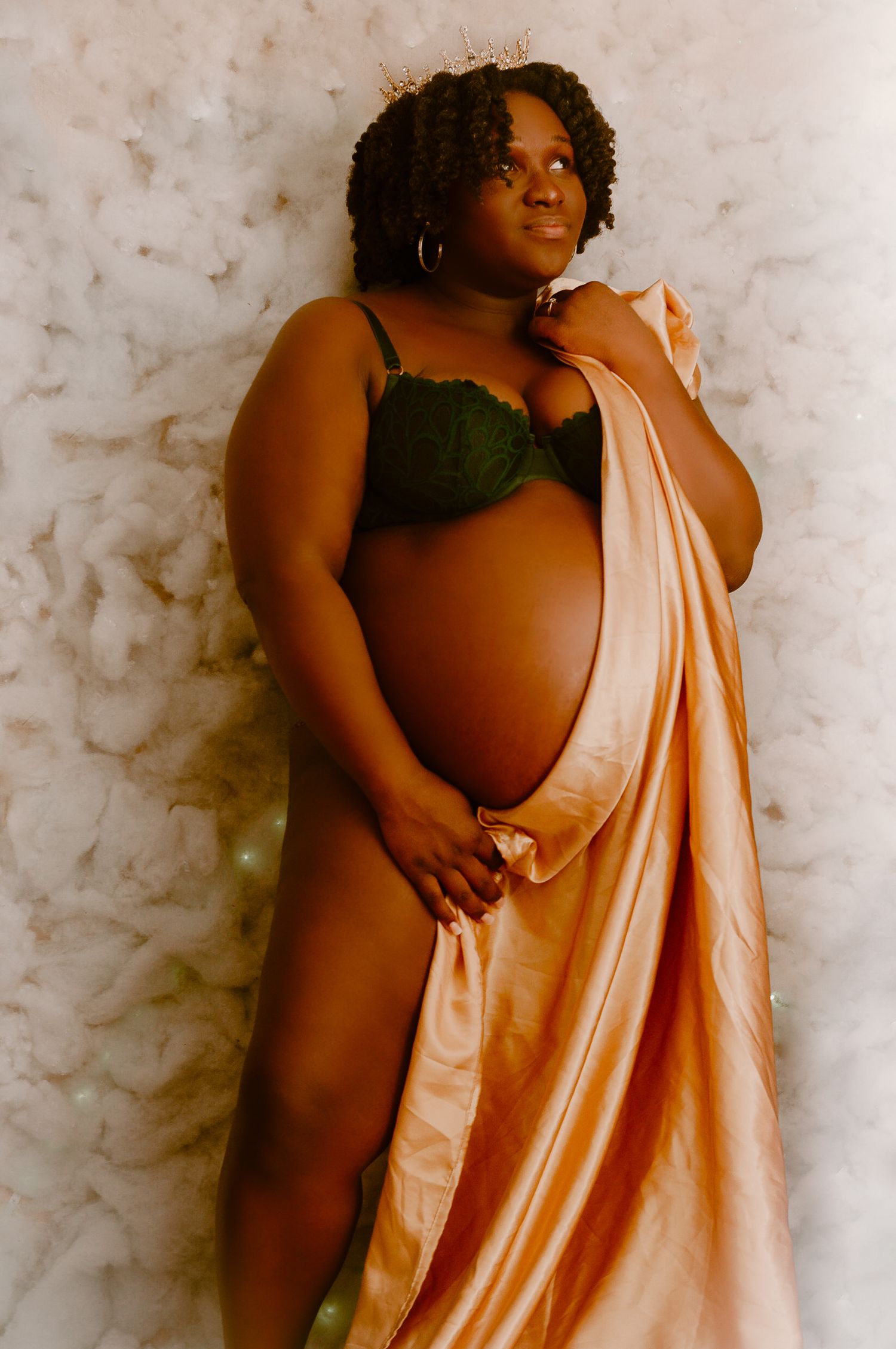 Pregnant Black woman in green lace bra draped in peach satin fabric, wearing a crown, posing for maternity portrait.