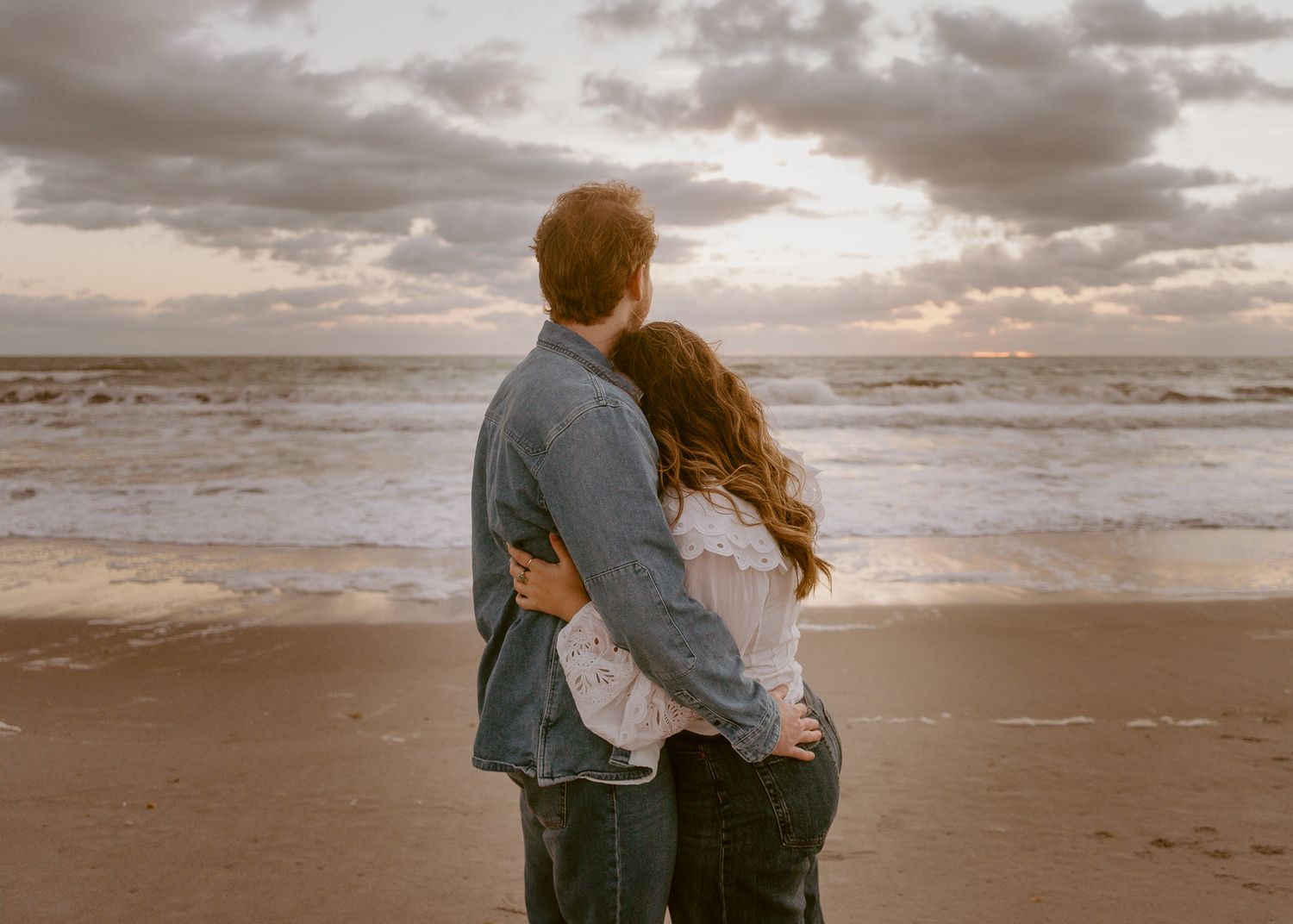 Couple embracing on a sandy beach at sunset with dramatic cloudy sky and ocean waves.