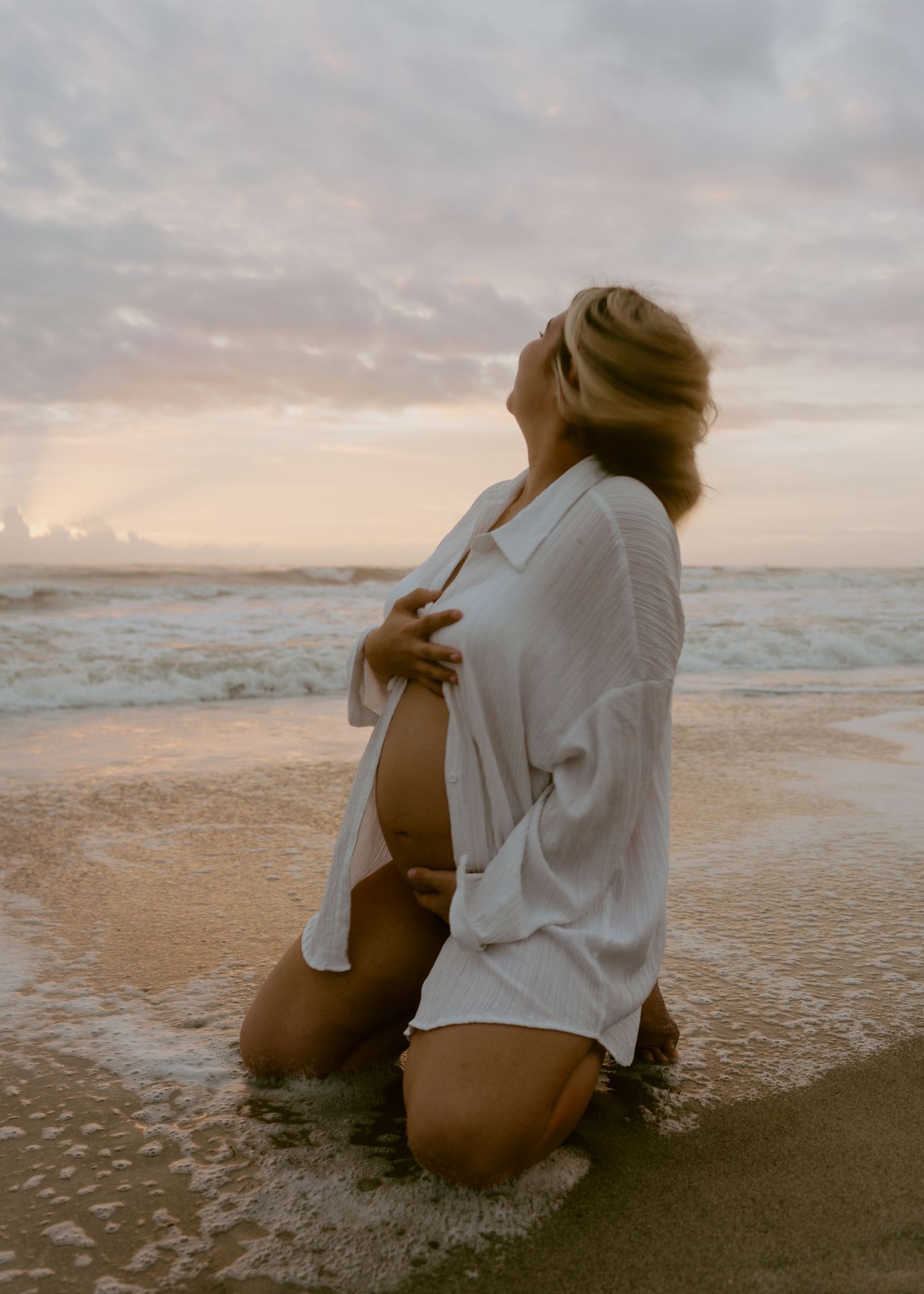 Pregnant woman in white shirt kneeling in ocean waves at sunset looking up at cloudy sky.