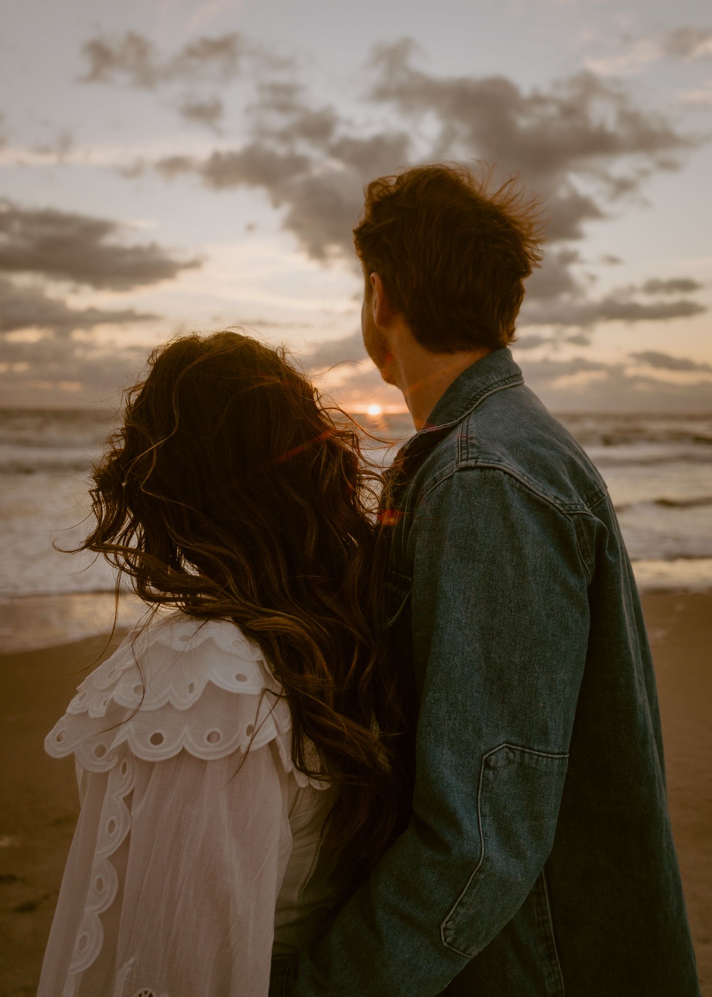 Couple standing together on a beach at sunset, woman in white dress with long curly hair, man in denim jacket.