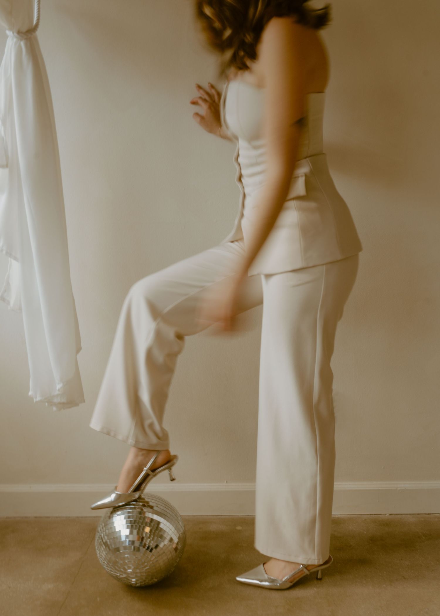Woman in cream satin pantsuit and silver heels stepping on a disco ball, posing elegantly indoors.