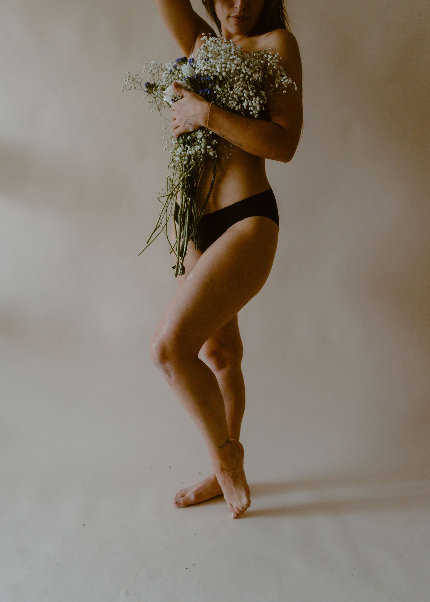 Woman in black underwear holding bouquet of white wildflowers, posing barefoot against beige studio backdrop.