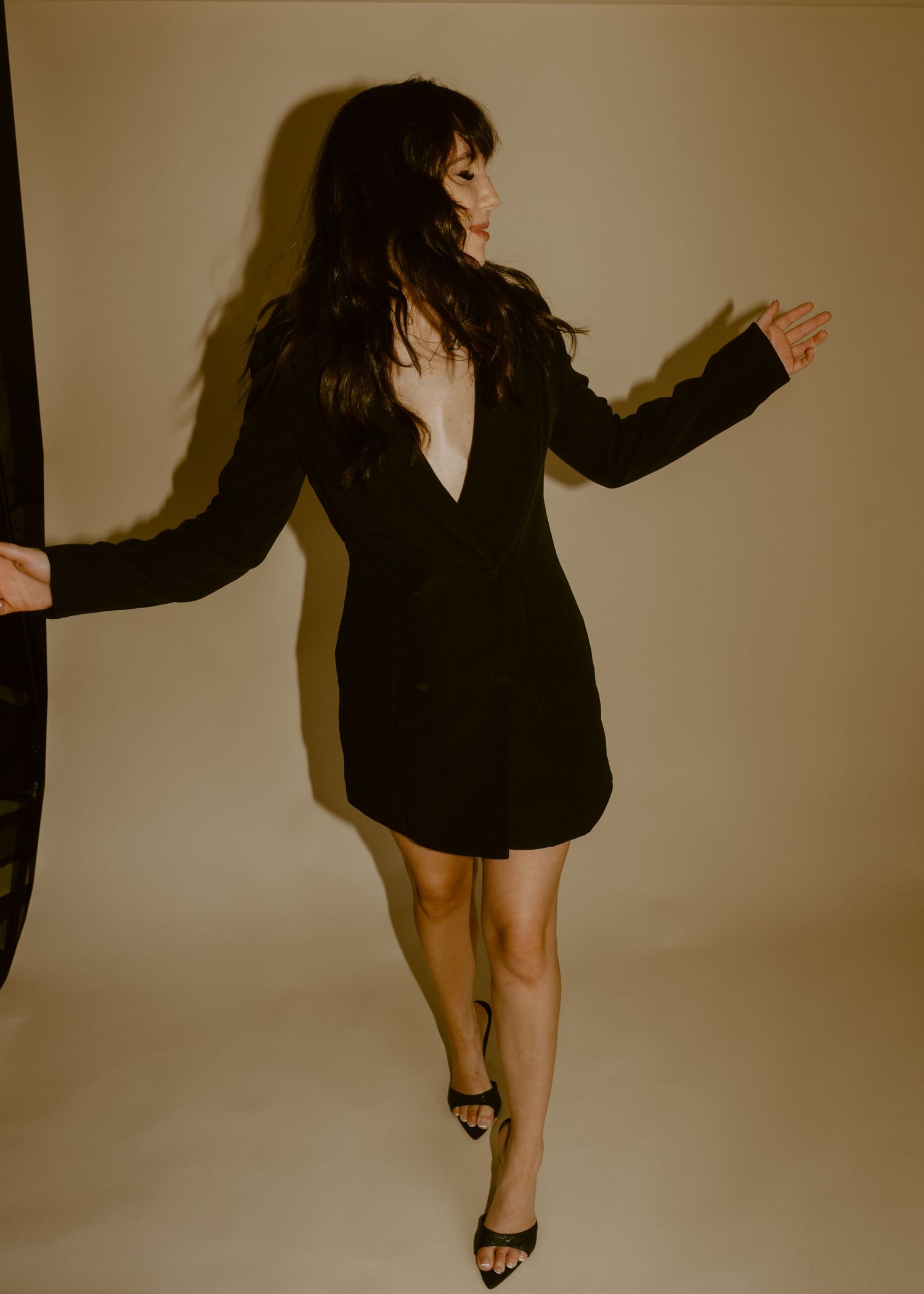 Woman in black plunge mini dress and heels poses against beige backdrop with dramatic lighting.