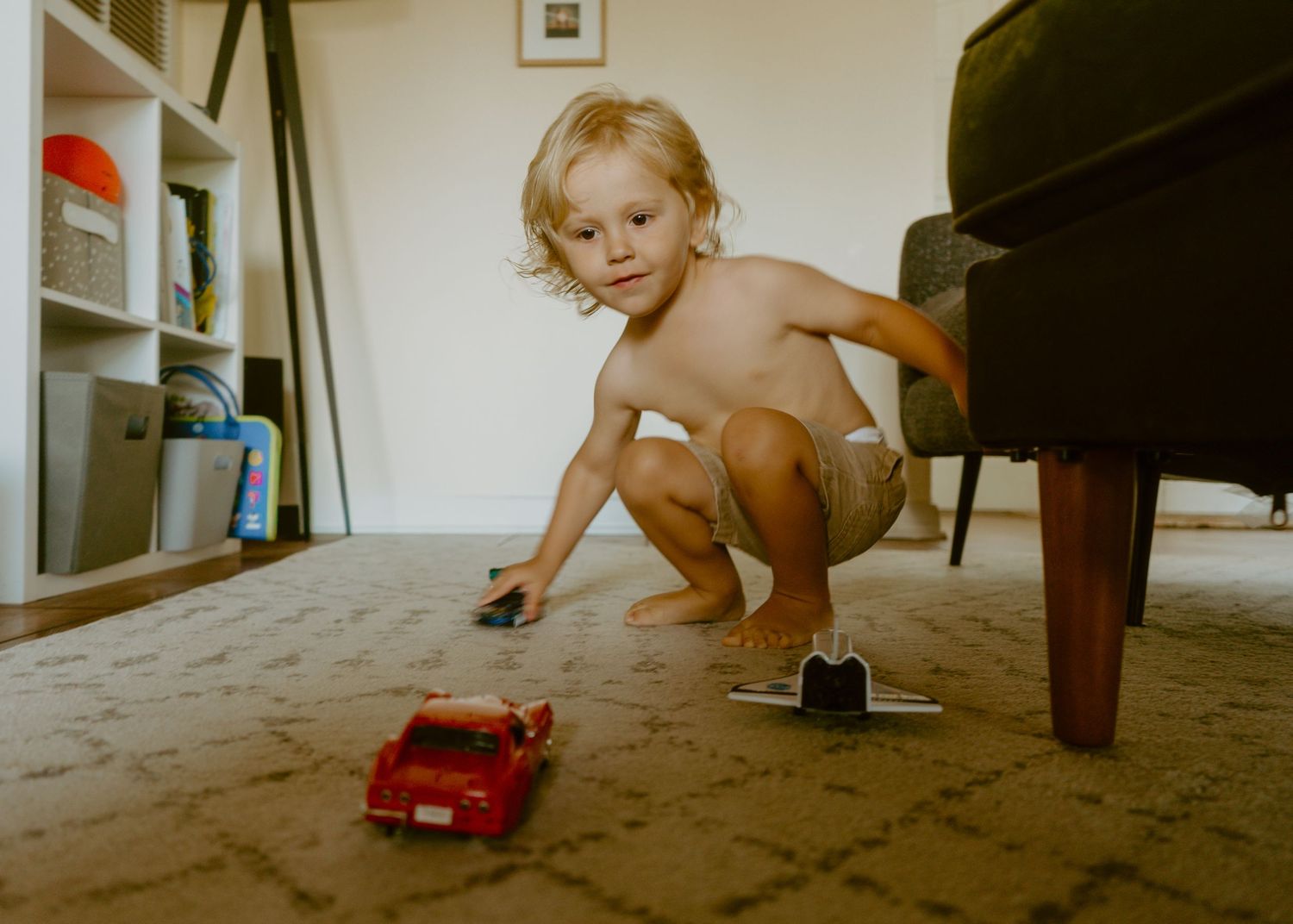 Young toddler playing with toy cars on carpet floor at home, crouching near sofa leg in living room.