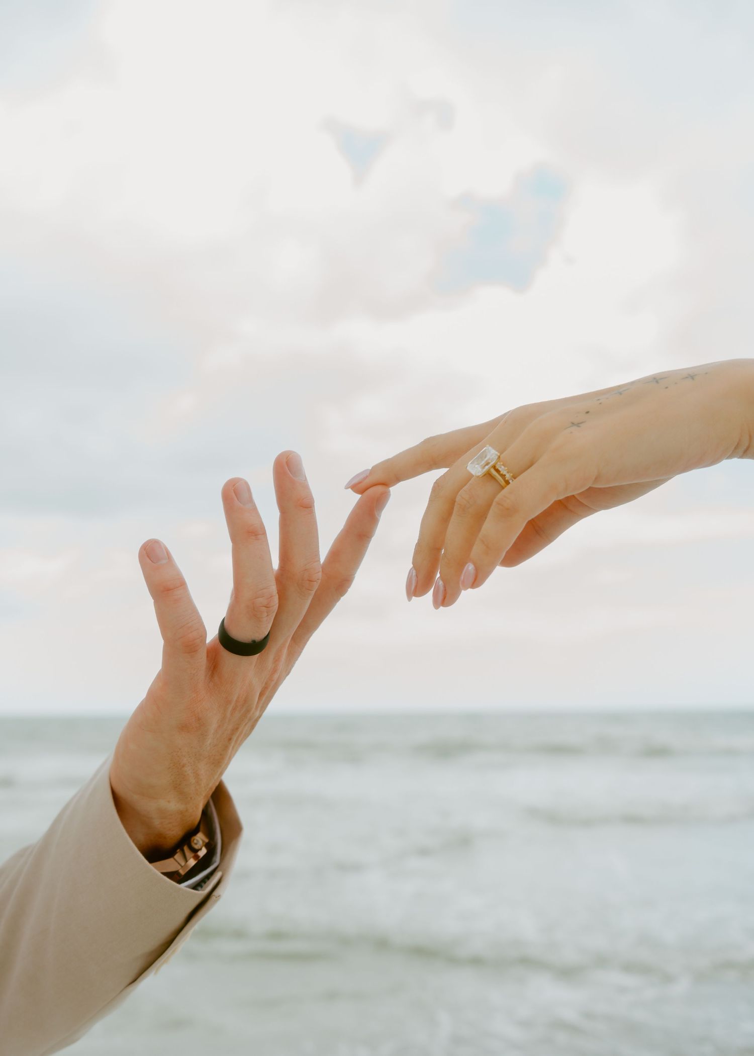 Two hands reaching toward each other at the beach, both wearing rings, against a cloudy sky backdrop.