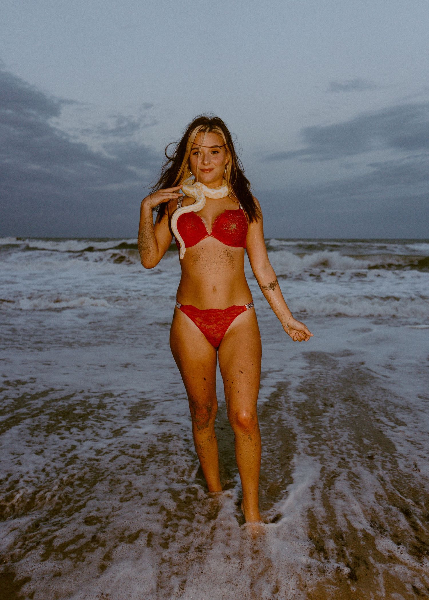 Woman in red bikini holding a white snake on a beach at dusk with dramatic cloudy skies.