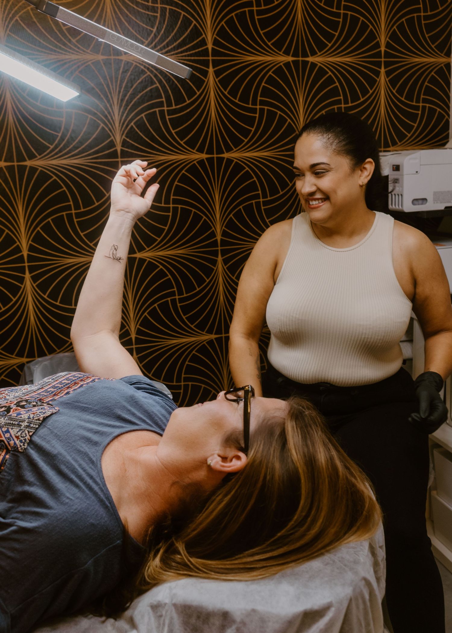 A smiling esthetician in black gloves stands over a relaxed client lying down in a modern salon with patterned walls.