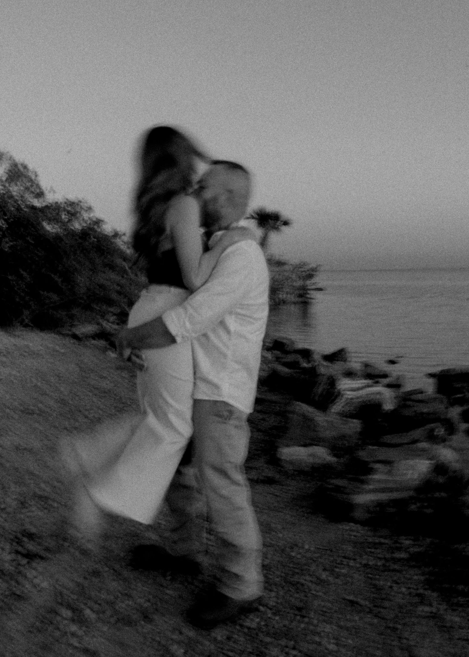 Blurry black and white photo of a couple embracing and kissing near a rocky shoreline at dusk.