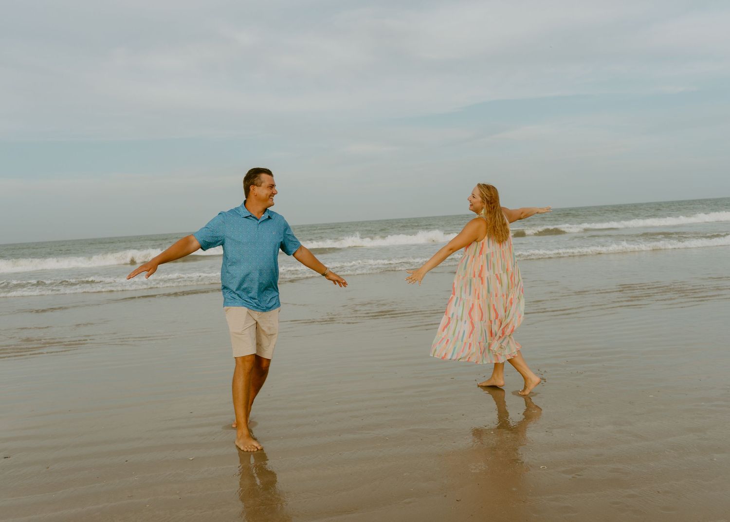 Couple holding hands and dancing playfully on a sandy beach with ocean waves in the background.