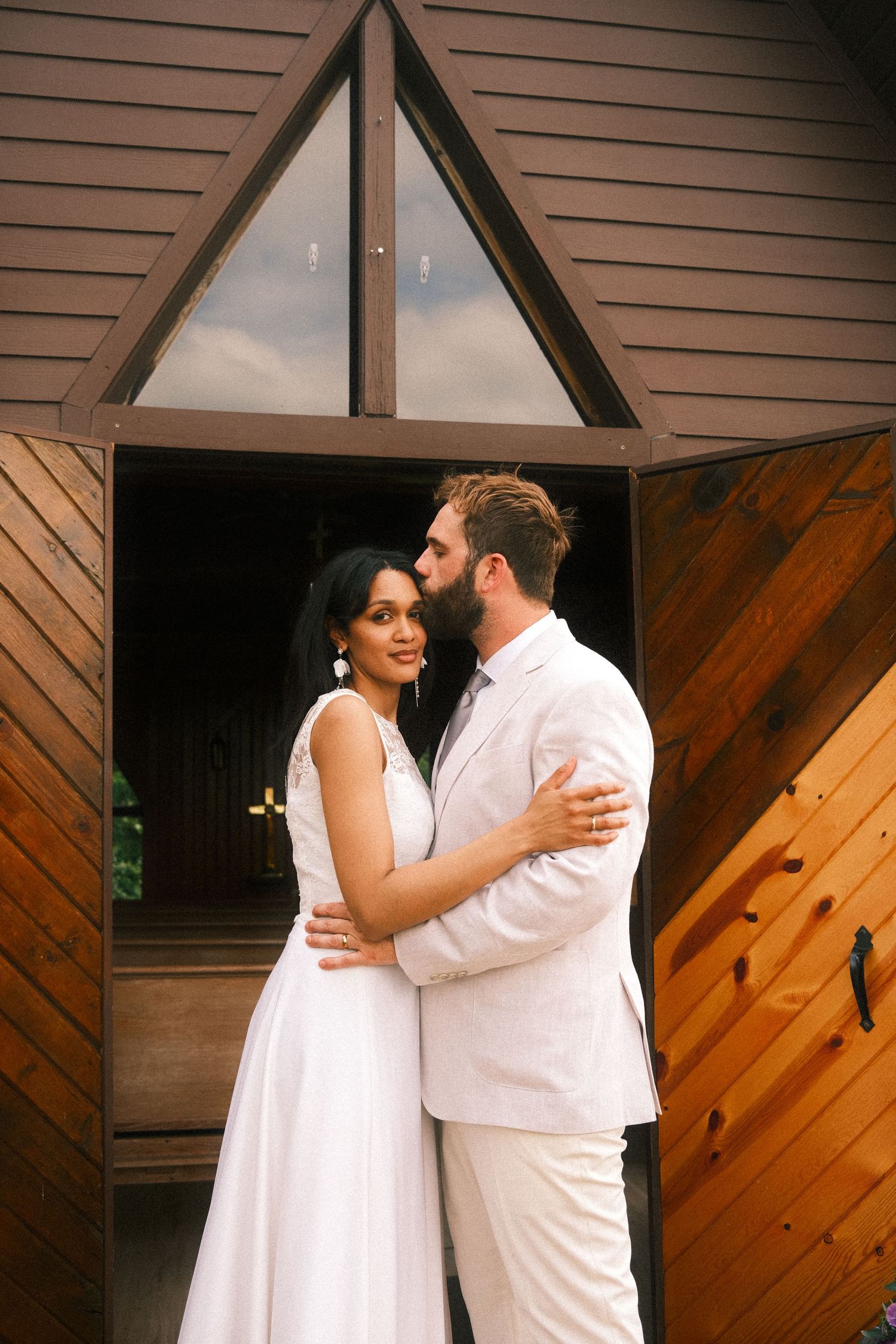 A couple dressed in white embraces romantically against a rustic wooden wall background during their wedding.