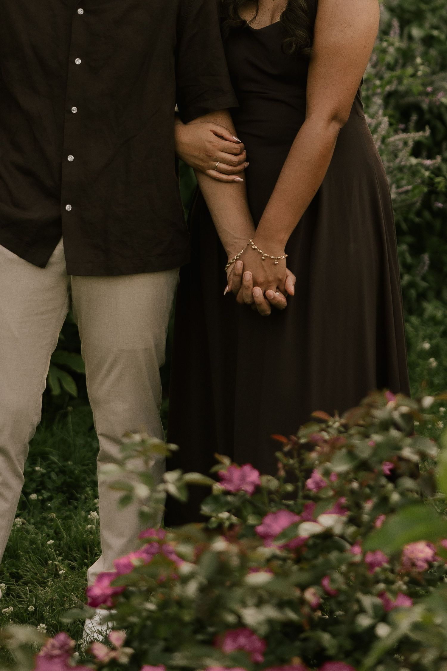 Couple holding hands in a garden with pink roses, woman wearing a gold hand chain bracelet and black dress.