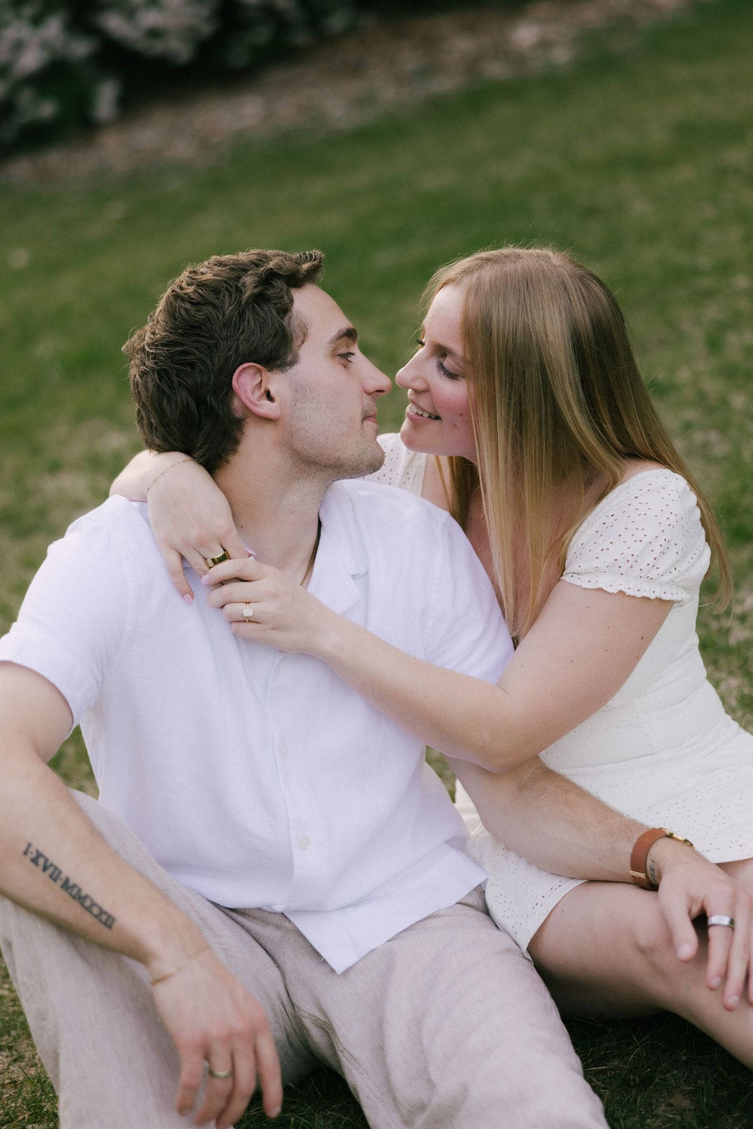 A couple sits on green grass, smiling at each other, the woman in white dress with arm around man in white shirt.