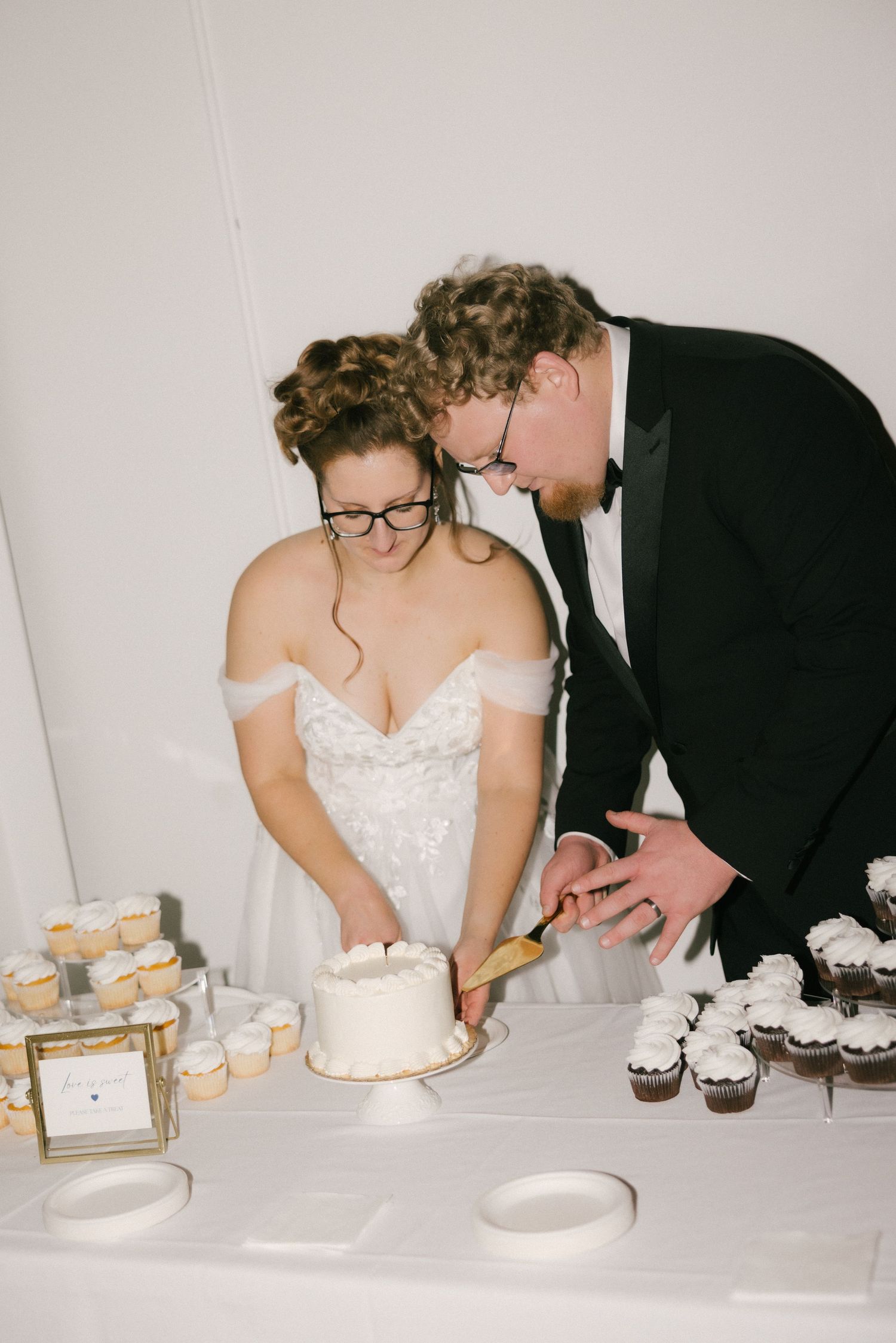 A couple in wedding attire cuts their white wedding cake together at the reception dessert table with cupcakes.