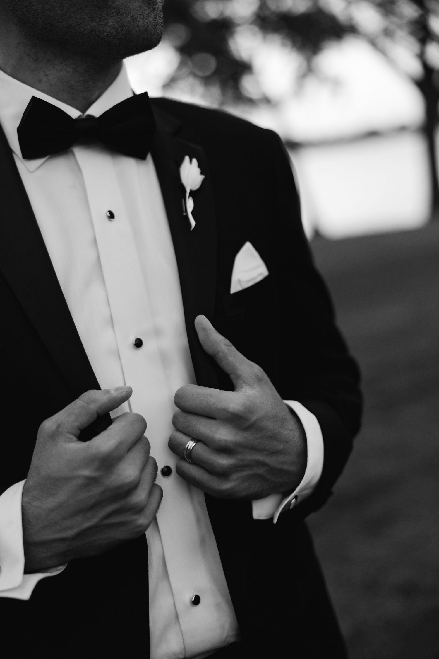 Black and white photo of a groom in a tuxedo with bow tie and boutonniere, adjusting his jacket on his wedding day.