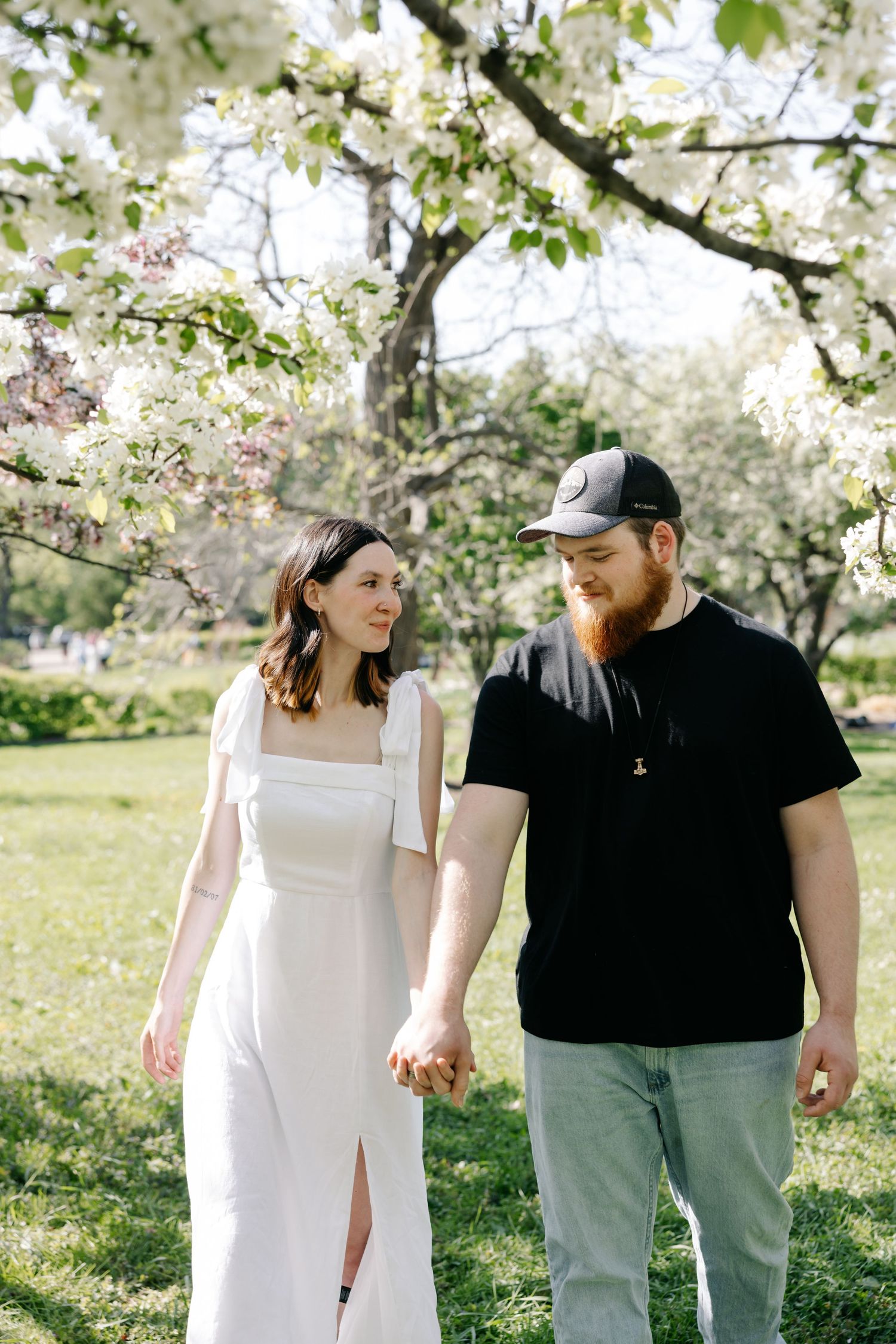 A couple holds hands while standing beneath blooming white cherry blossom trees in a spring garden setting.
