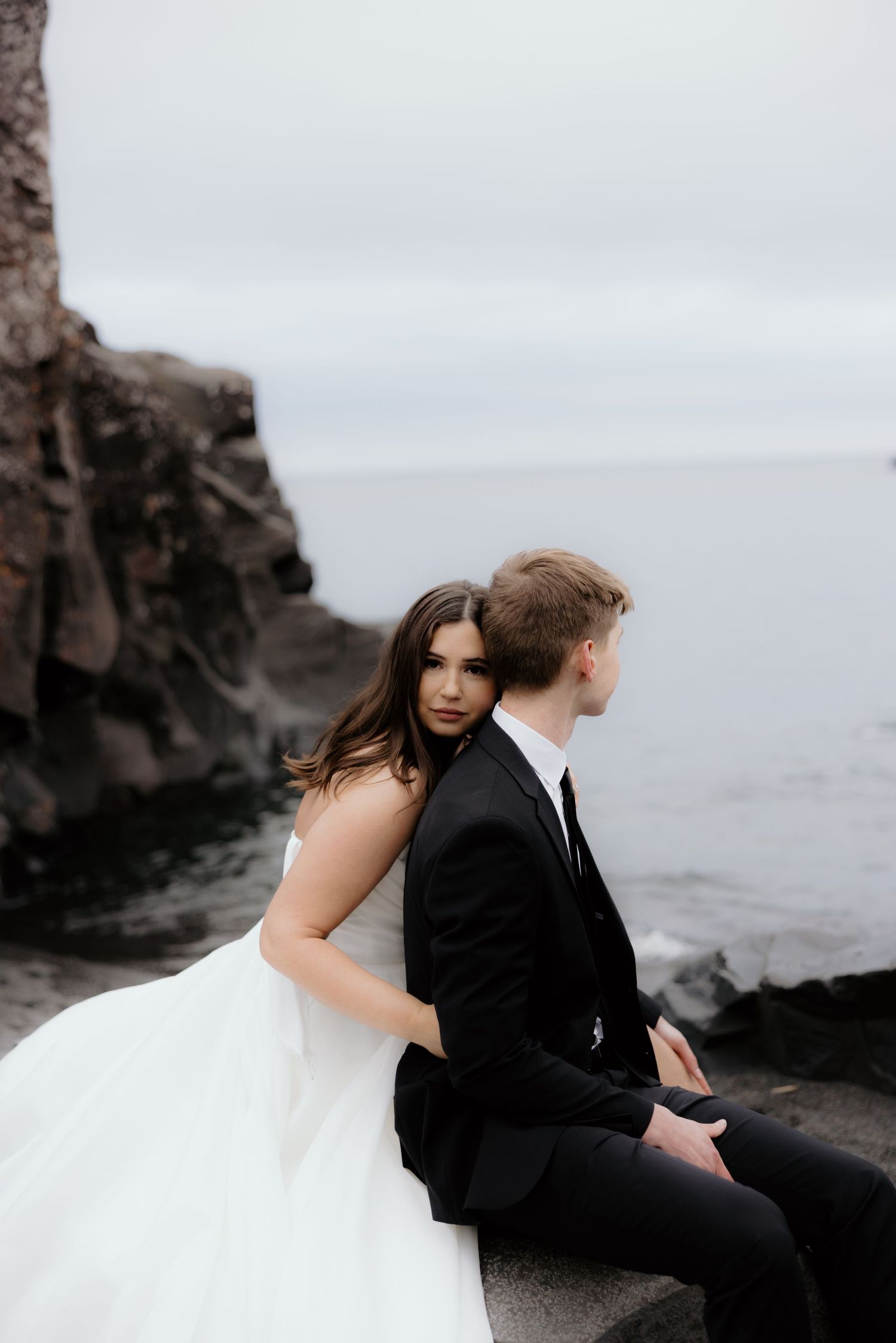 A couple in wedding attire sits on rocky cliffs overlooking a misty ocean on their wedding day.