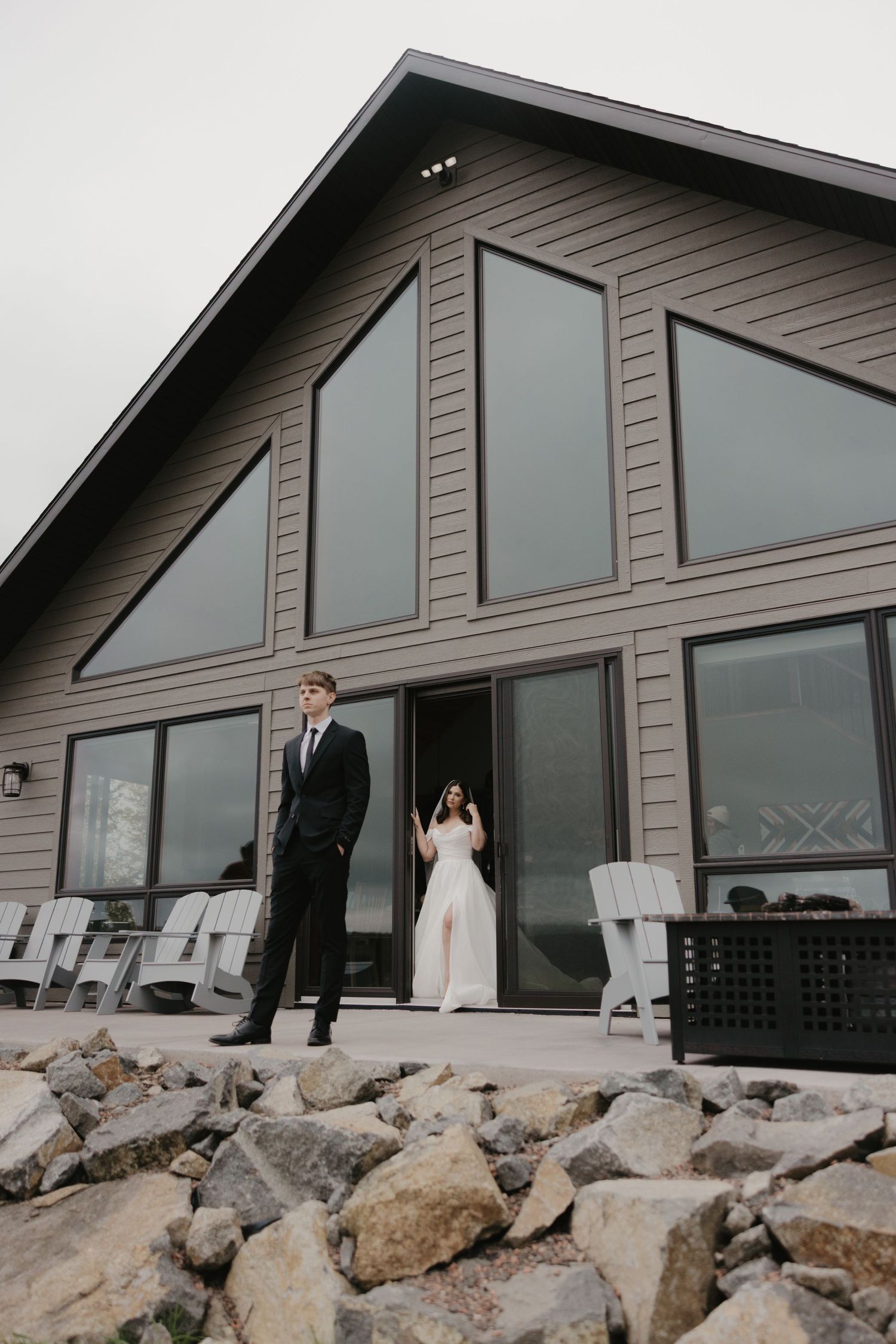 A modern A-frame house with large windows and stone foundation features a couple standing in formal attire on the deck.