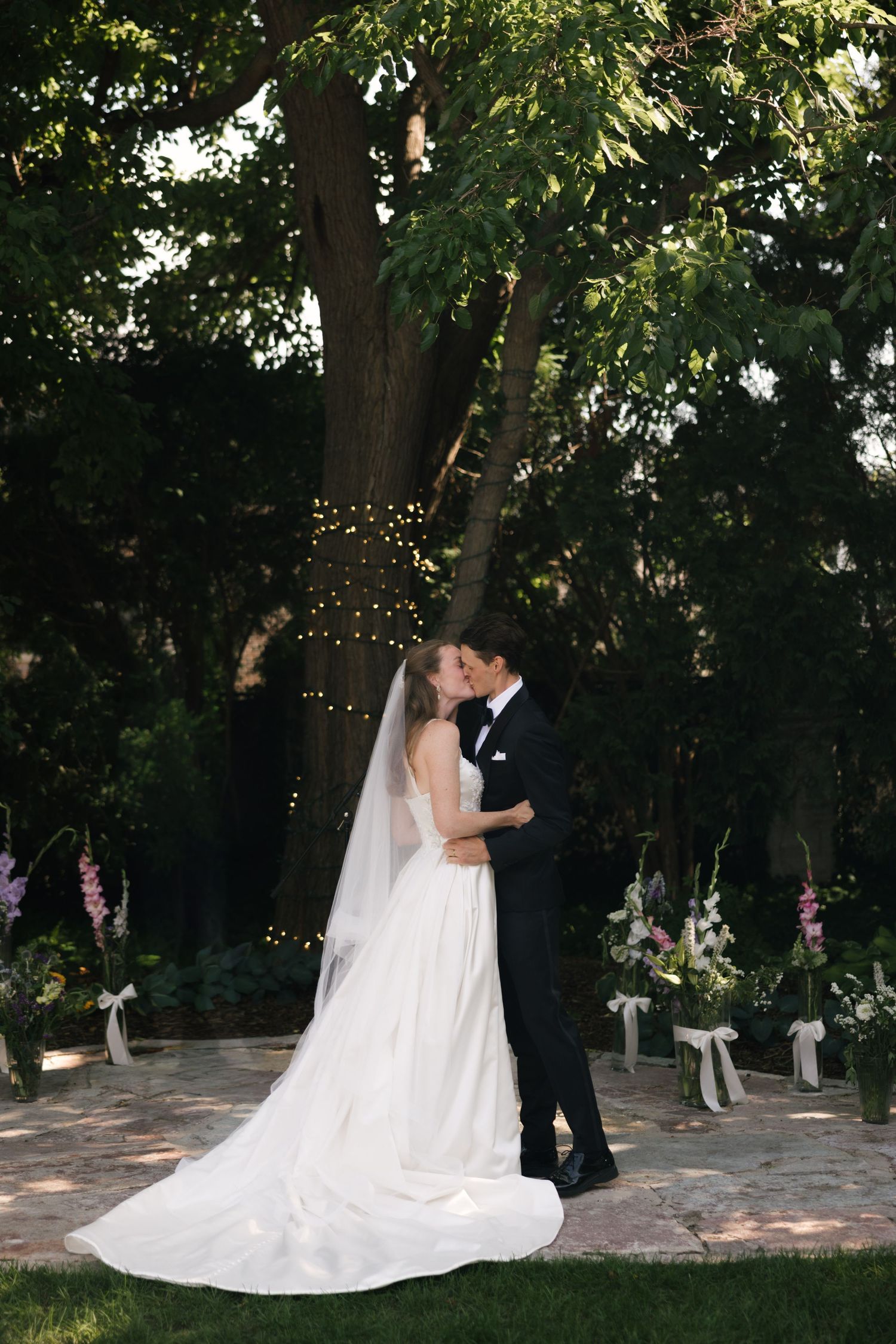 Bride and groom share first kiss under a large tree with fairy lights at an outdoor garden wedding ceremony.