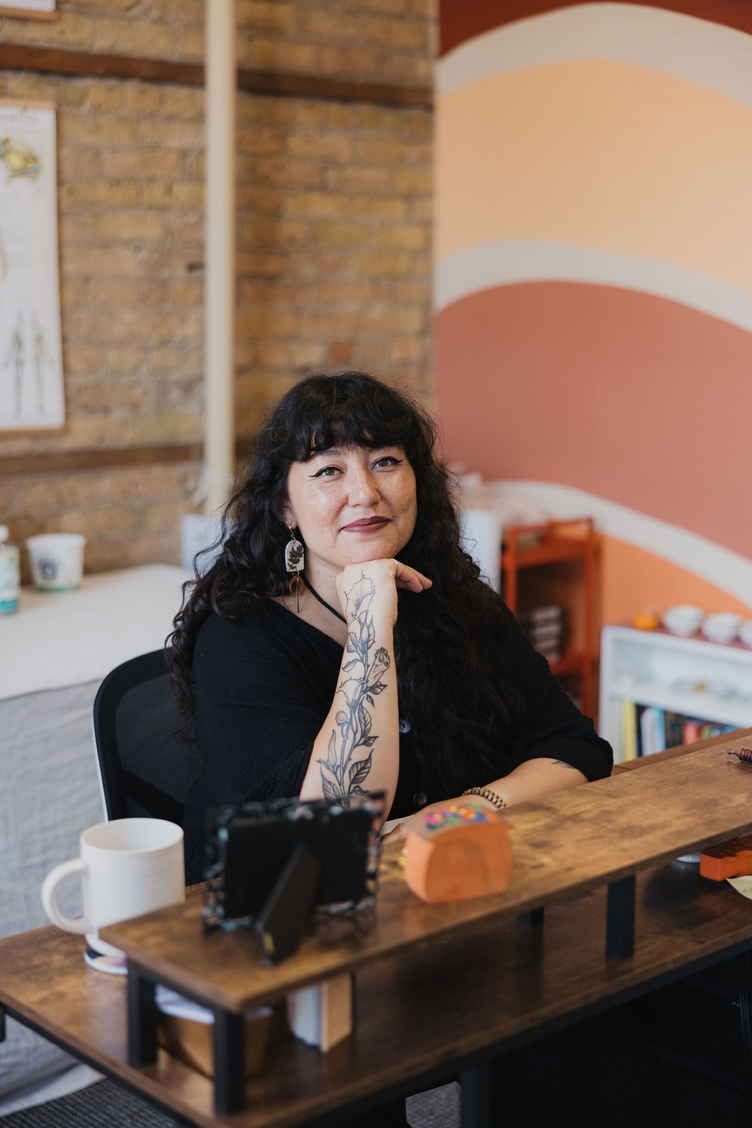 Woman with dark bangs and tattoo sleeves sits at wooden table in cafe with exposed brick wall and orange accents.