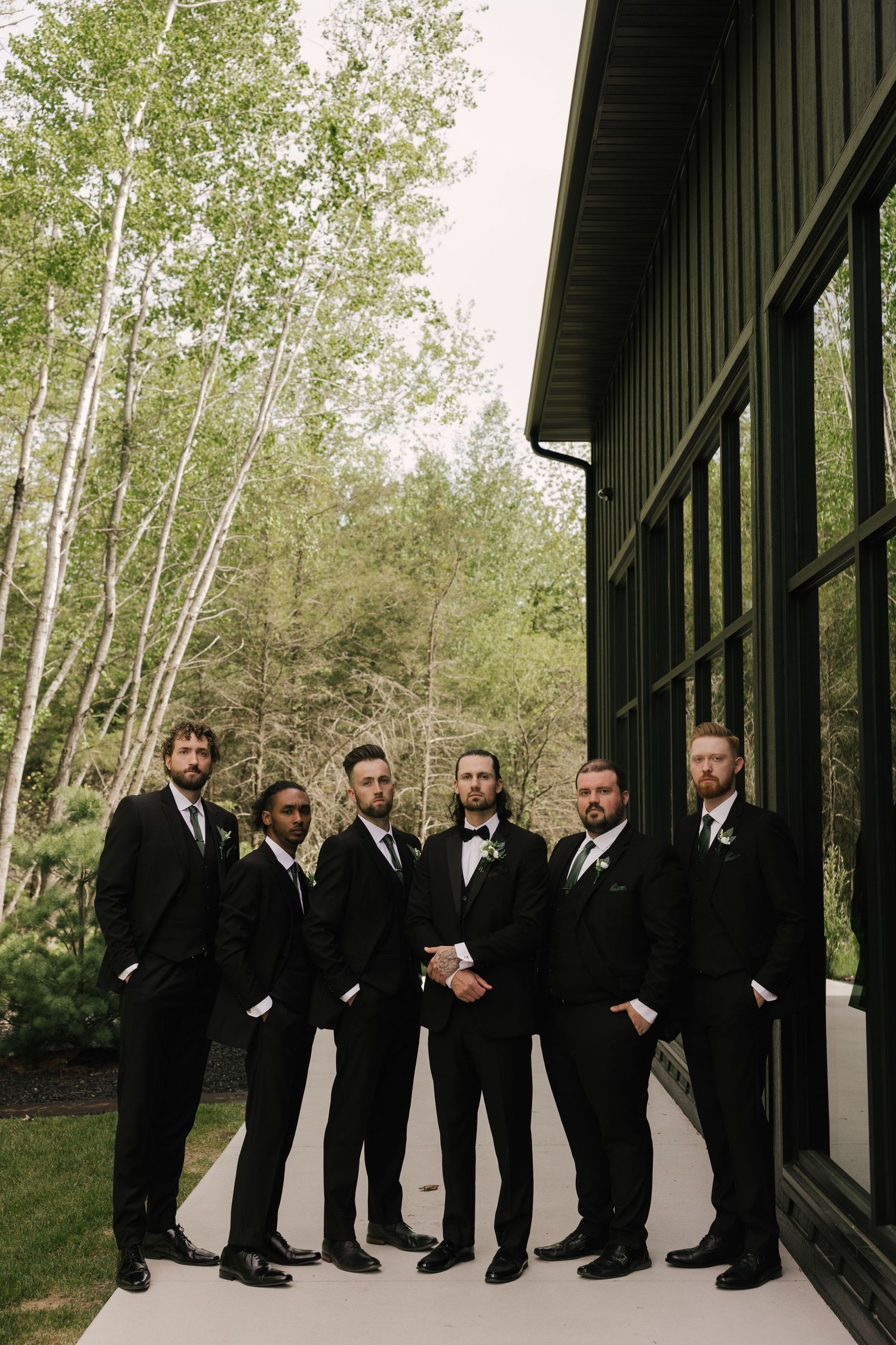 Six groomsmen in black tuxedos pose outside a modern dark green building surrounded by birch trees.