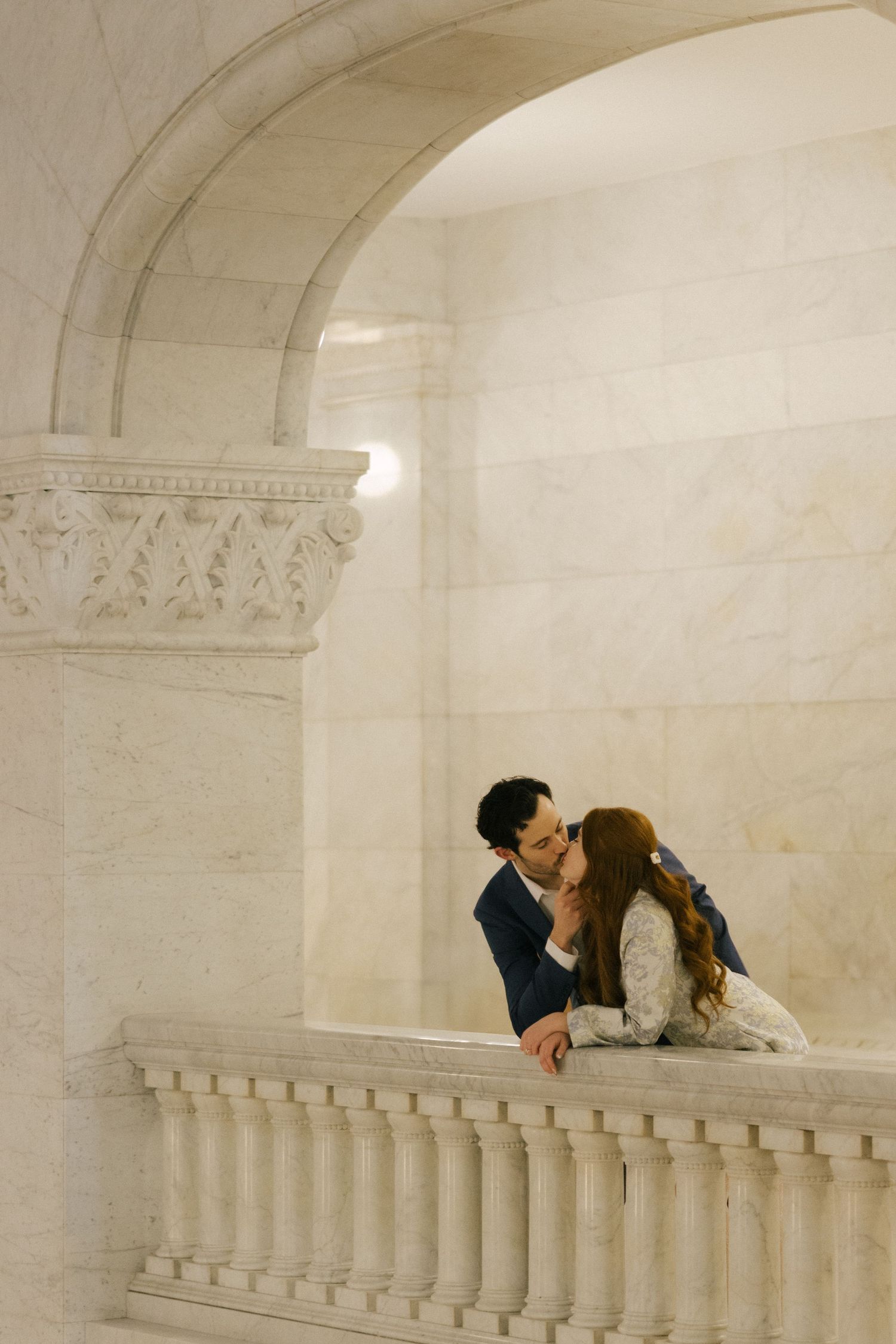 Two people share an intimate moment in a white marble architectural space with ornate balustrades and columns.