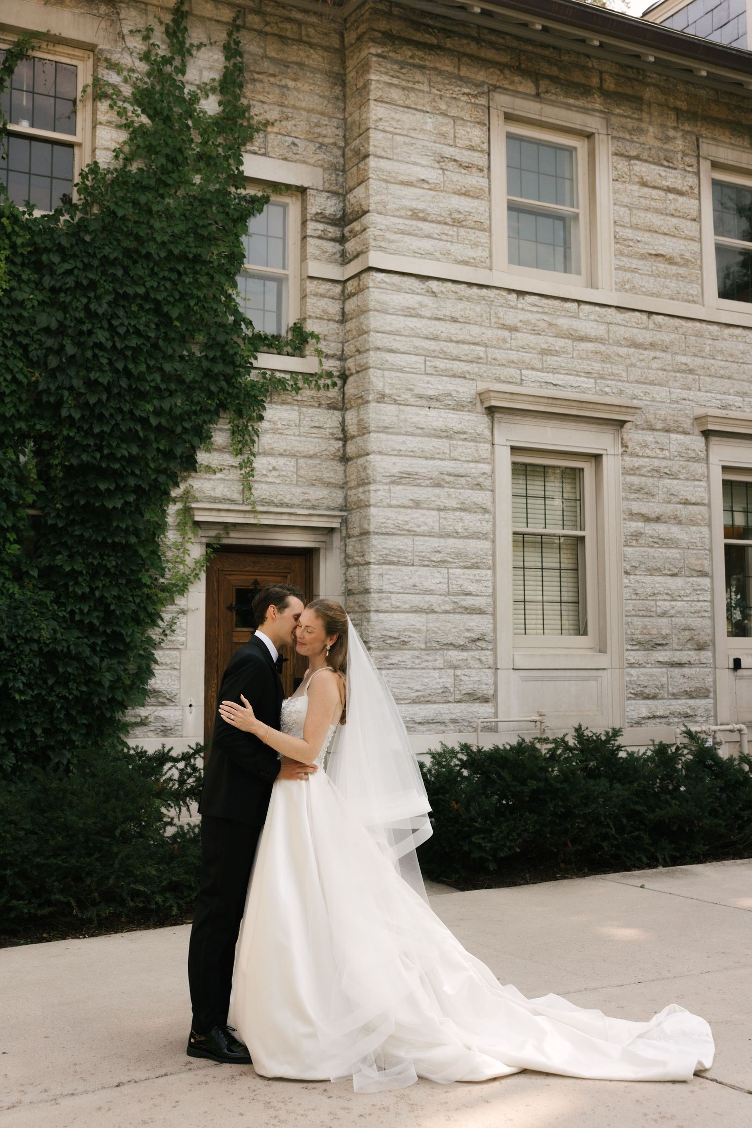 A bride and groom share a romantic kiss outside a stone building covered in ivy on their wedding day.