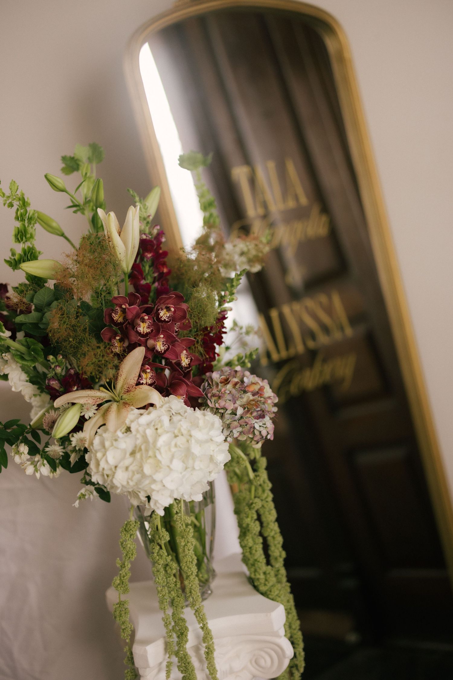 Elegant wedding floral arrangement with white hydrangeas, burgundy orchids, and greenery beside a gold mirror.