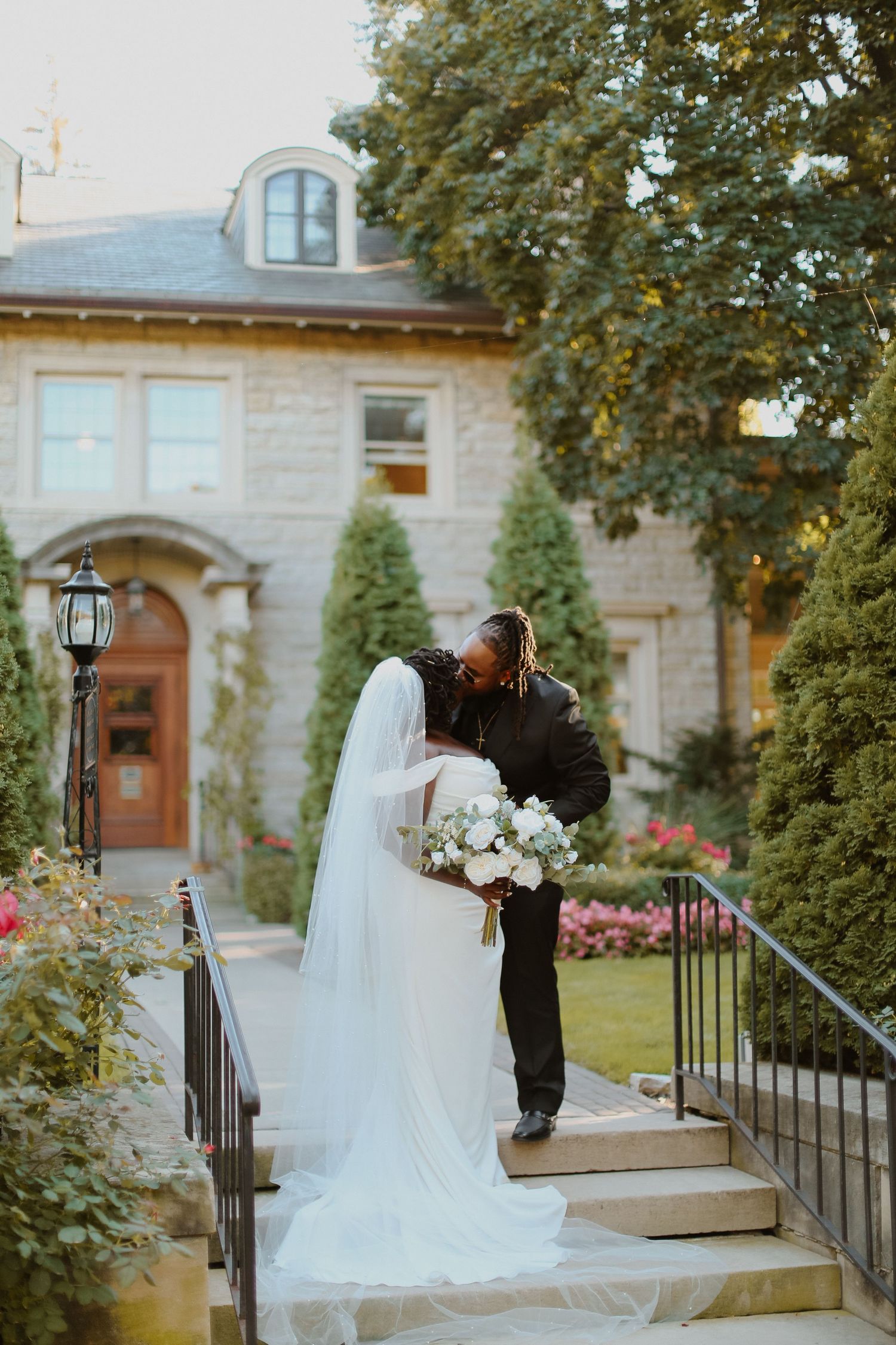 A couple shares an intimate moment on stone steps outside a historic mansion with a flowing white veil and bouquet.