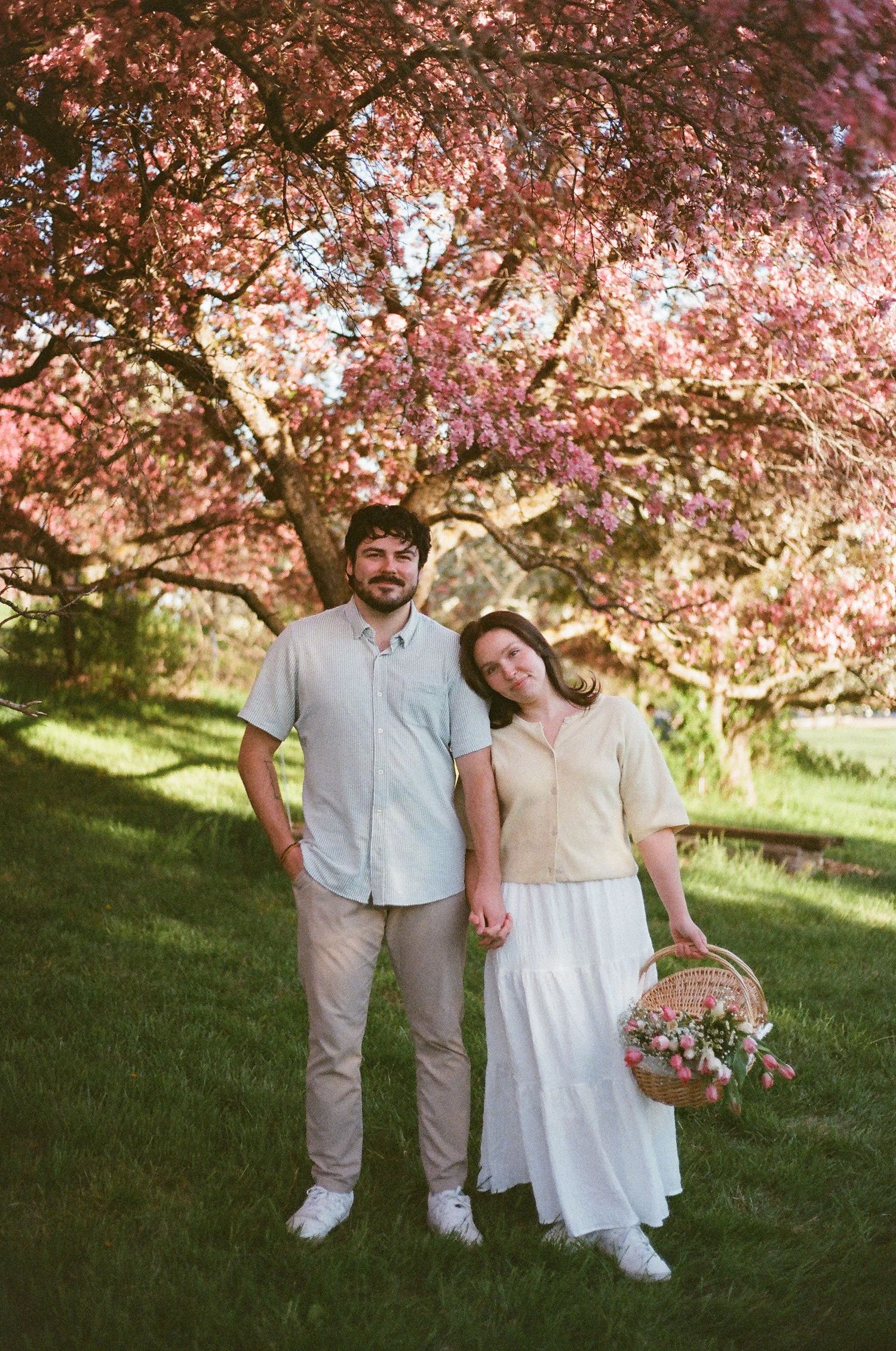 Couple holding hands under pink cherry blossom tree, woman in white skirt holding flower basket, spring garden setting.