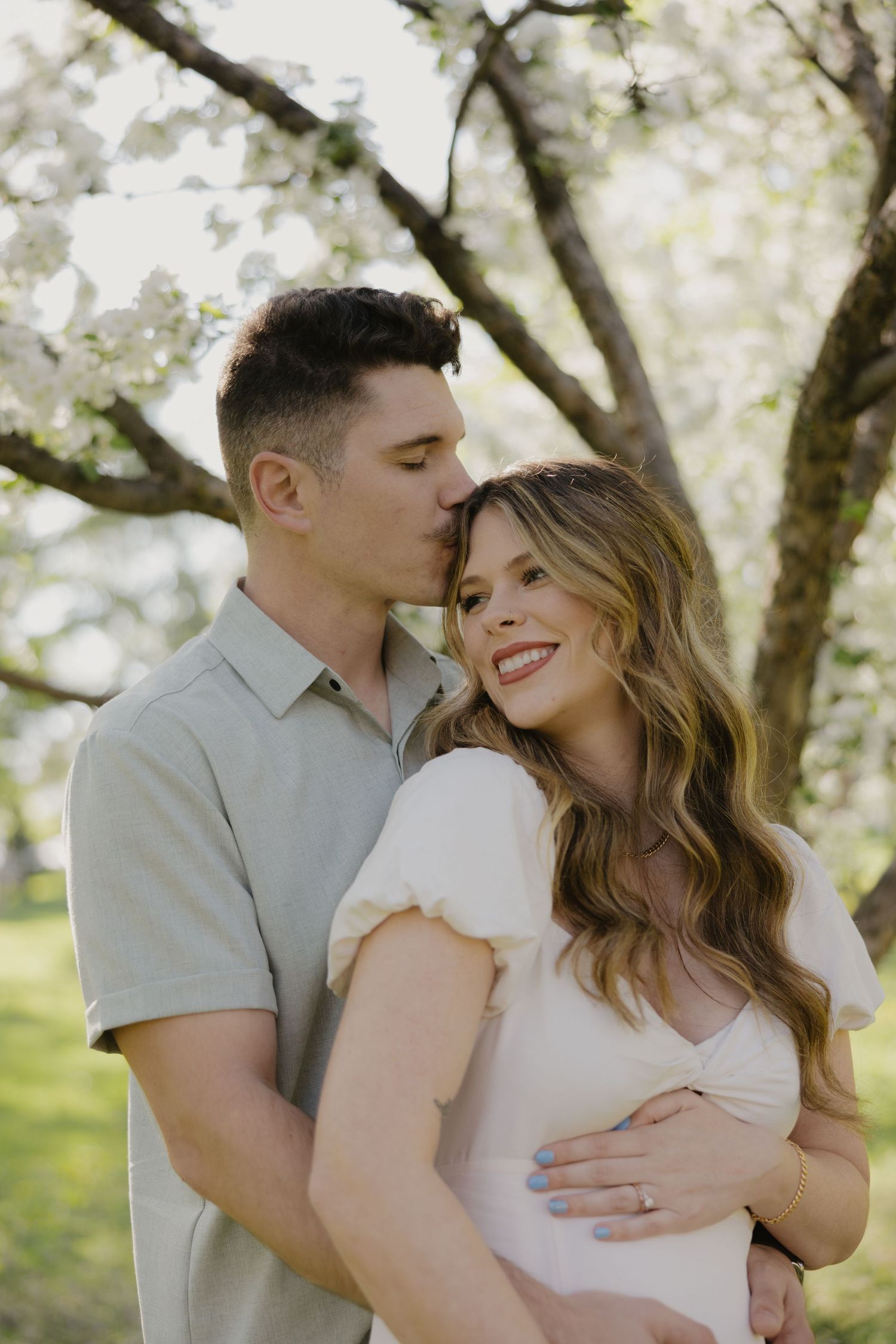A romantic couple embraces outdoors during springtime with blooming trees in soft natural lighting.
