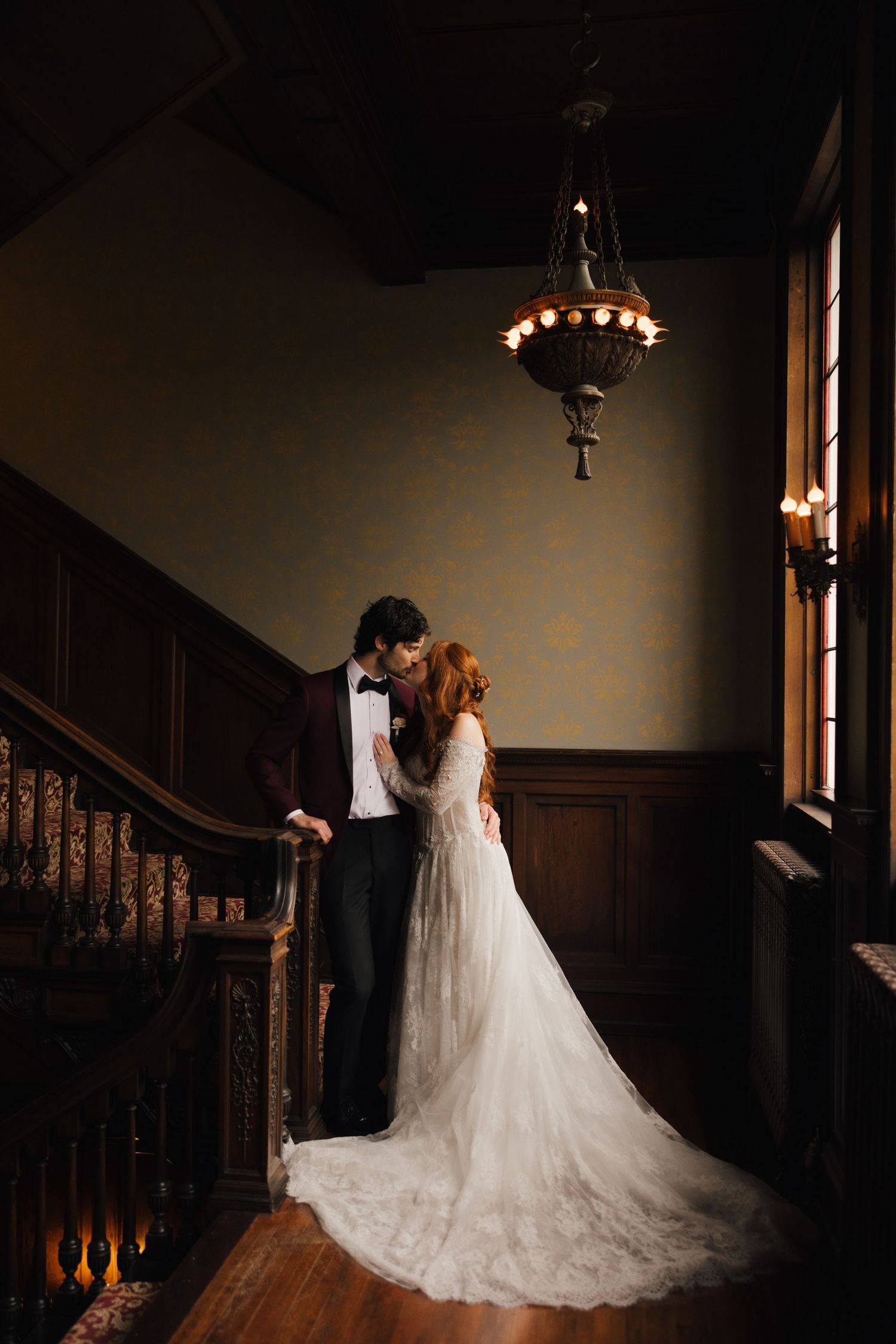 Bride and groom share a romantic moment on an ornate wooden staircase beneath a vintage chandelier.