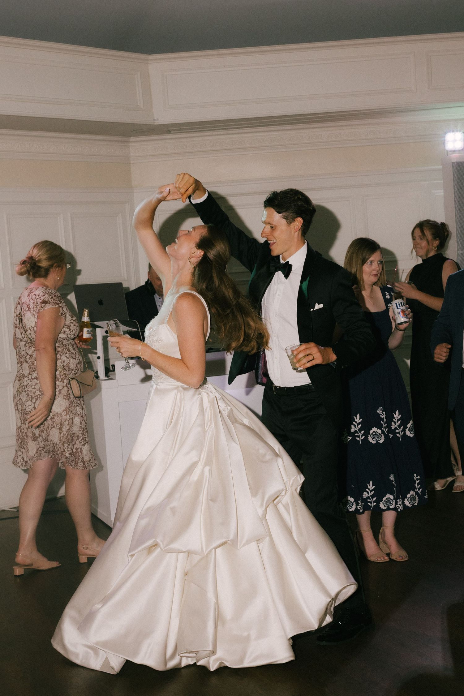 Bride and groom share a joyful first dance at their wedding reception, guests celebrating in the background.