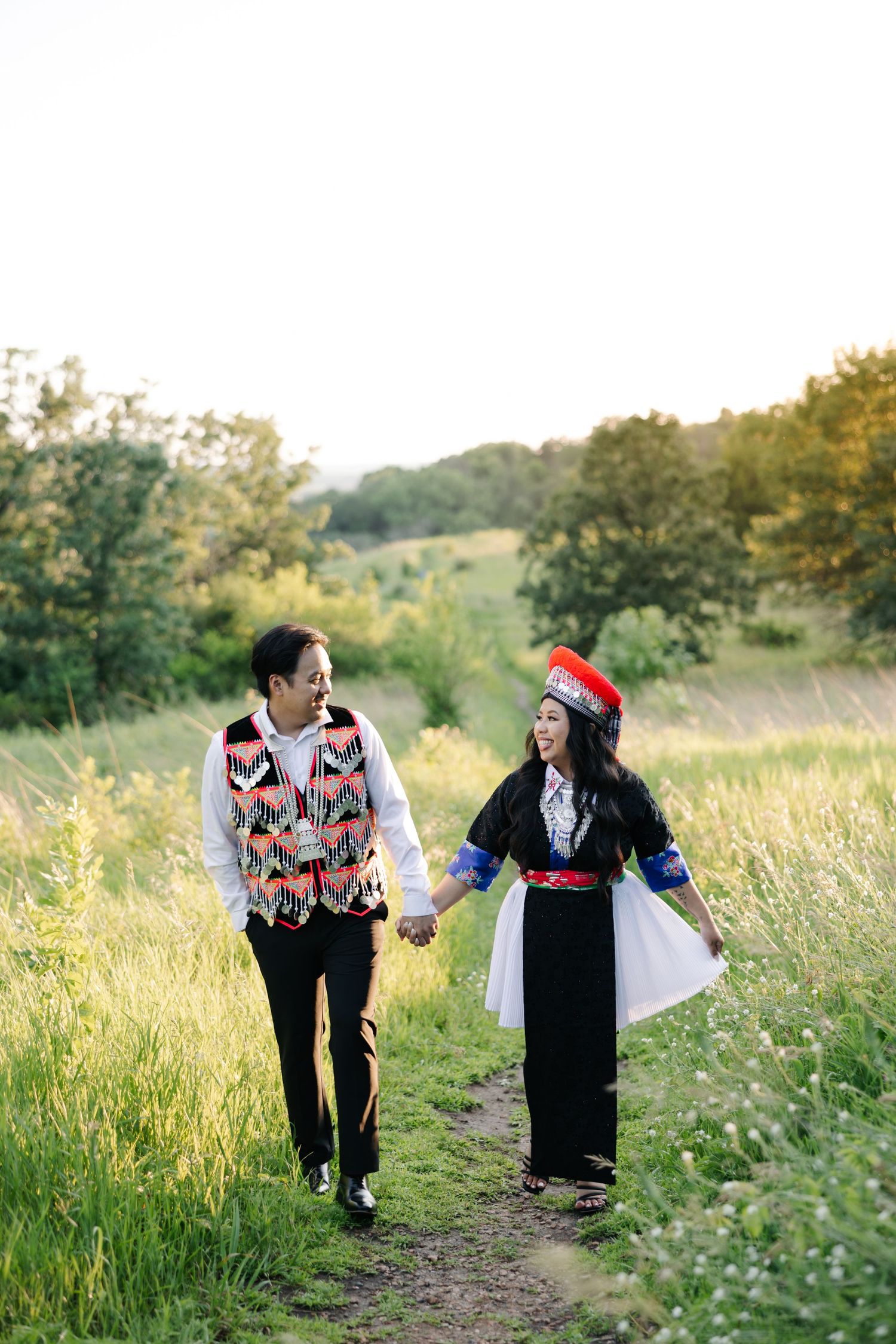 A couple in traditional folk costumes walk hand-in-hand along a nature trail through a meadow at sunset.