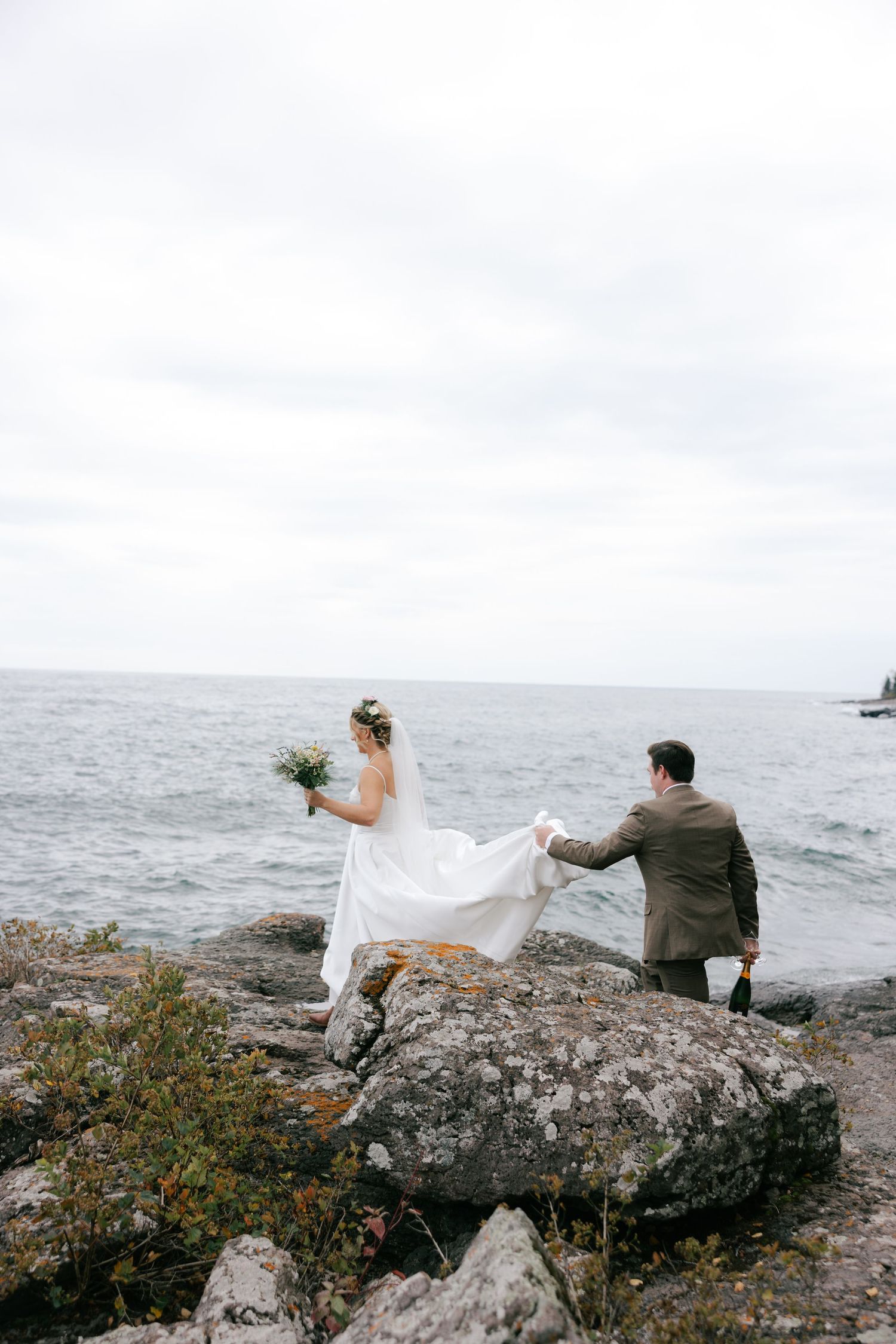 A couple sits on coastal rocks overlooking the ocean as wind blows a white veil in a romantic seaside moment.