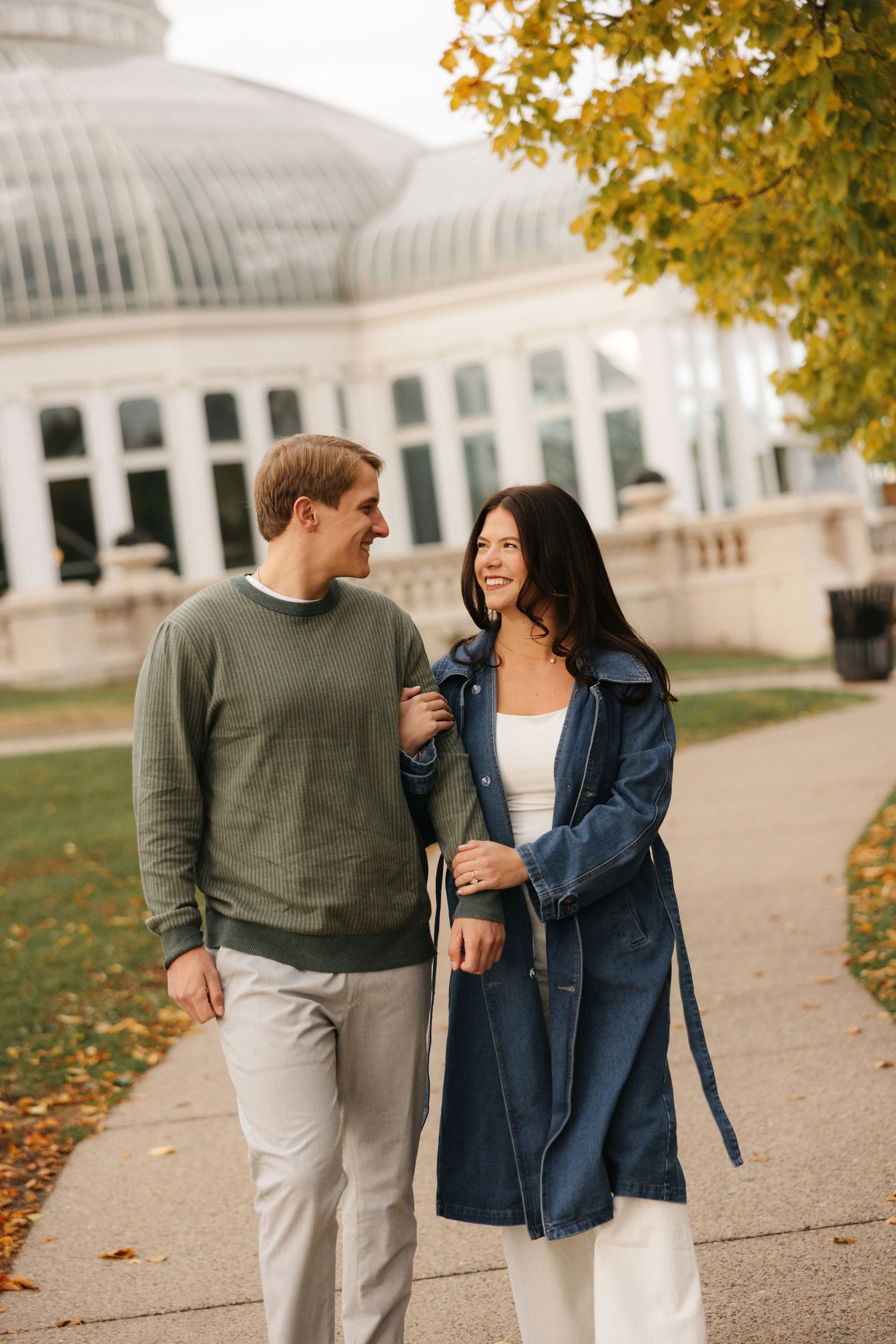 A couple walking together in autumn near a white conservatory building with golden leaves on trees.