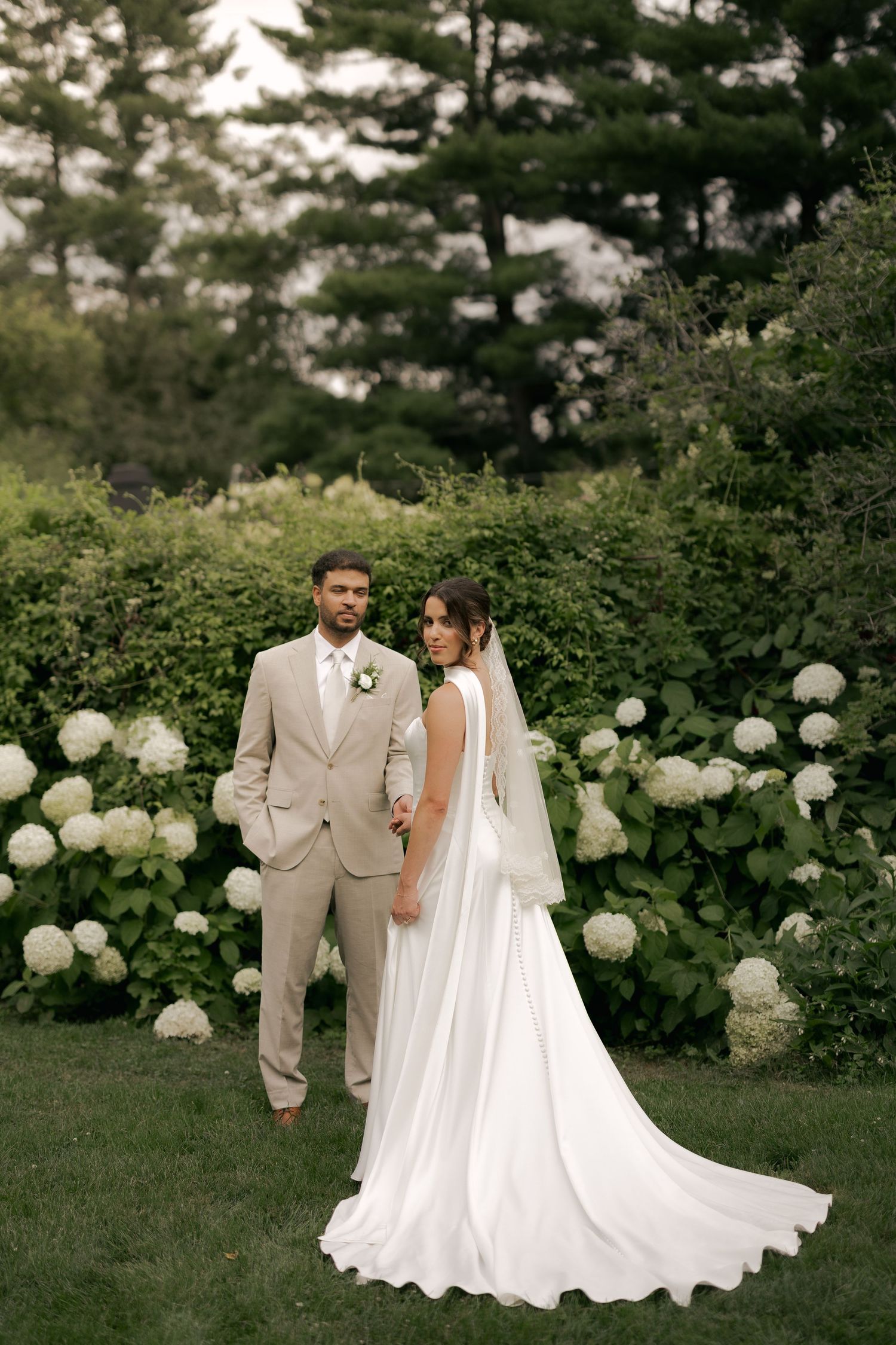 Bride in white gown with veil and groom in tan suit pose before white hydrangeas in a lush garden setting.