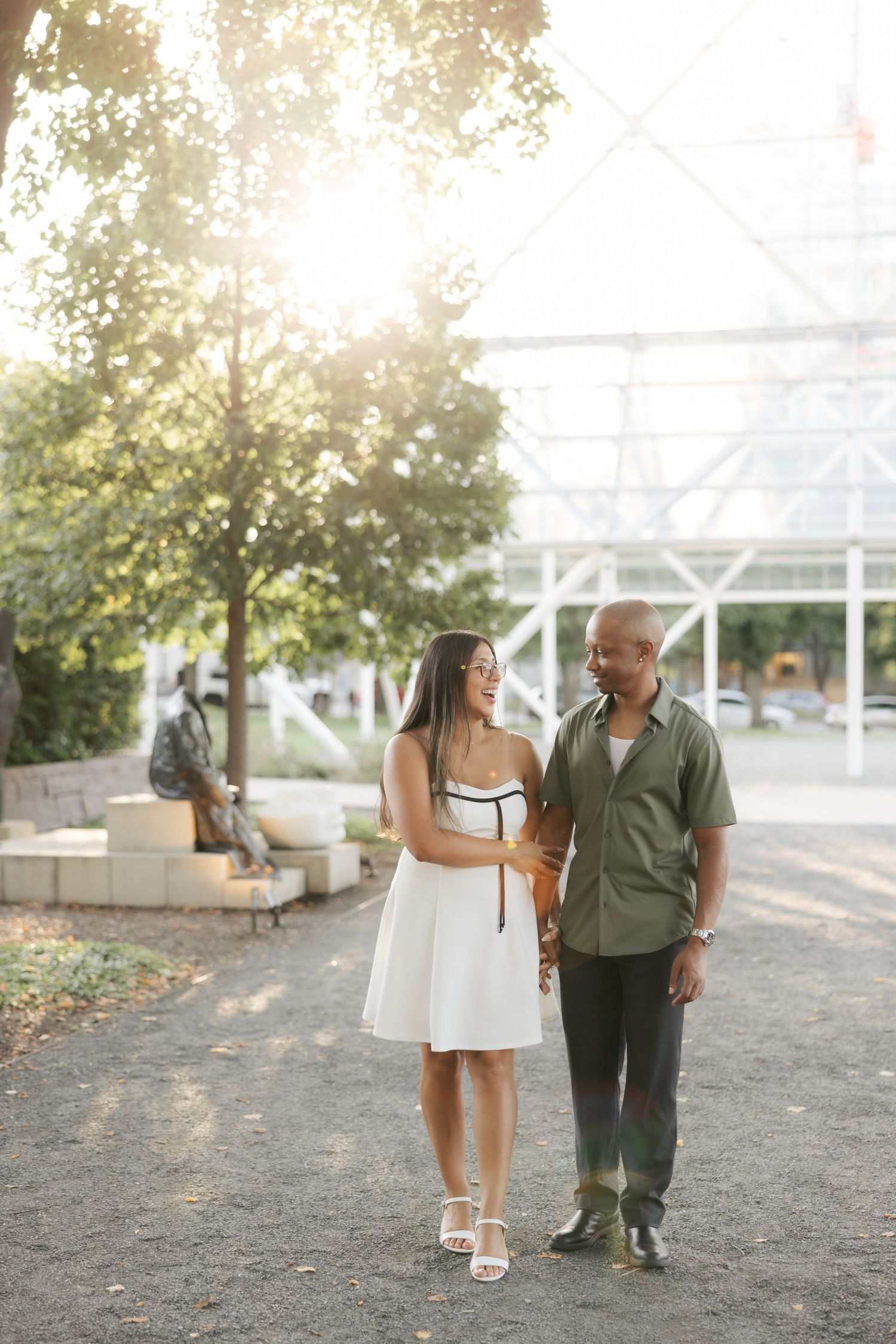 A couple walks together on a gravel path near a white greenhouse structure on a sunny summer day.